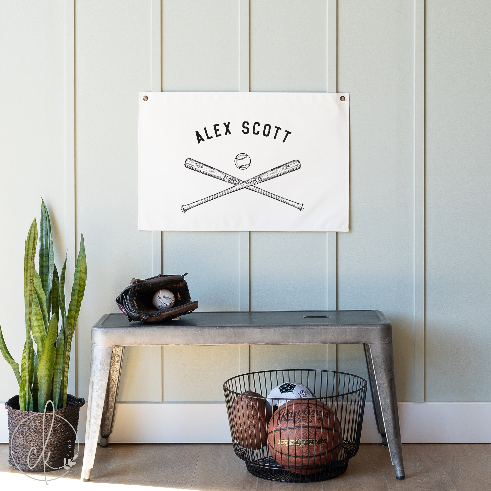 A personalized canvas flag with Alex Scott written above a sketch of crossed baseball bats and a baseball, hanging on a light blue paneled wall. Below the flag is a metal bench with a baseball glove and ball on top.