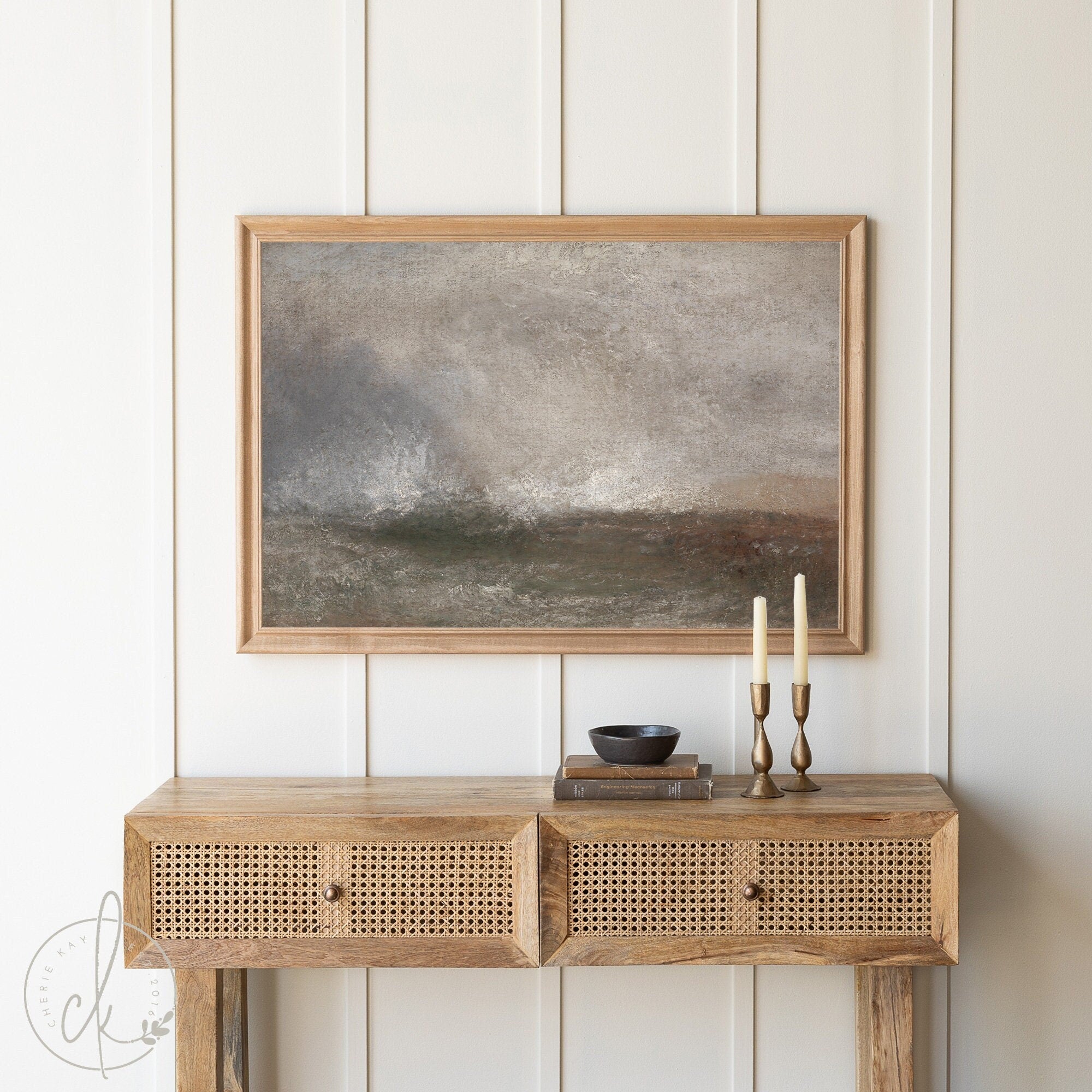 Framed abstract landscape painting in neutral tones, displayed above a rustic wooden console table with two brass candle holders and a small black bowl, set against a light paneled wall.