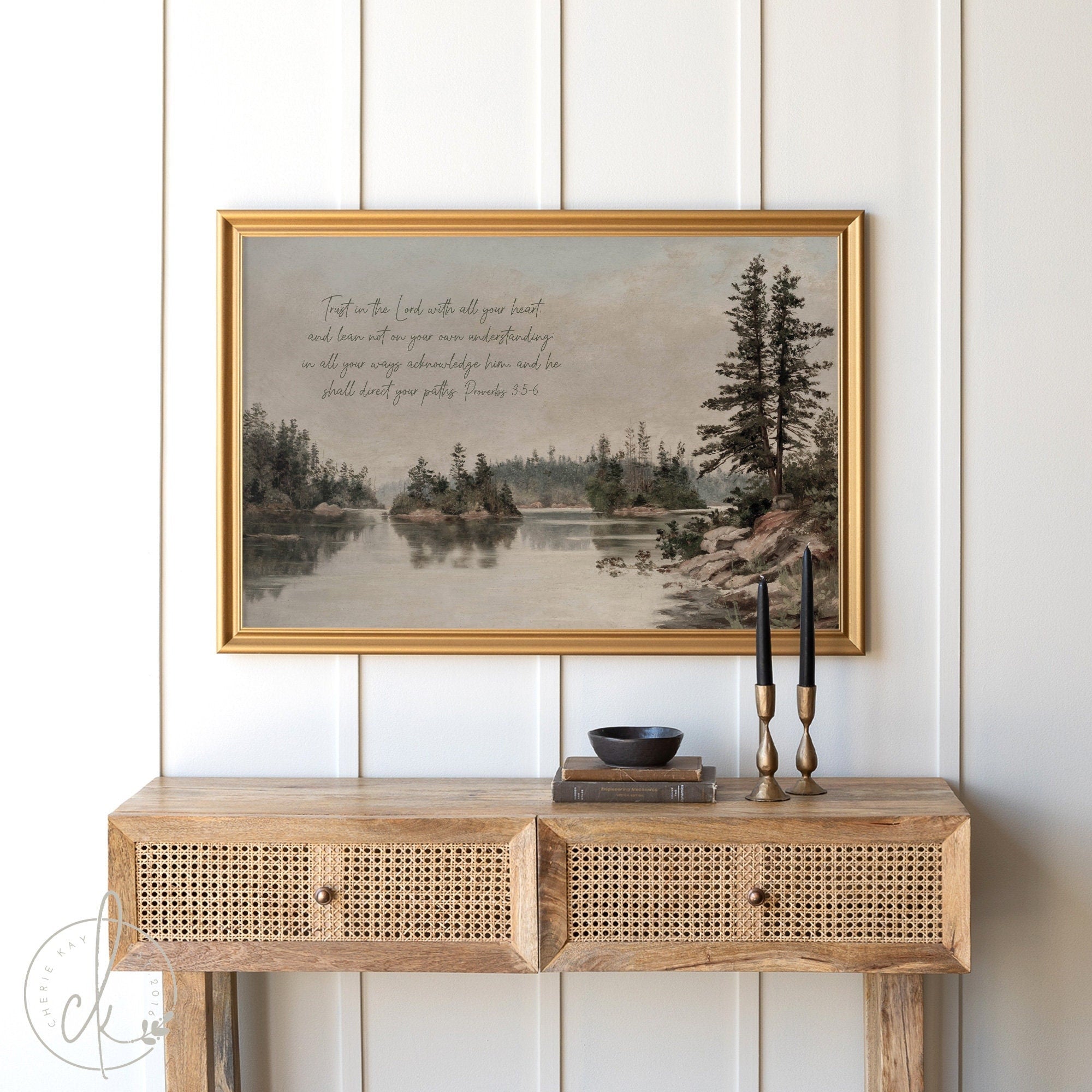 Vintage framed artwork of a tranquil landscape scene featuring a lake, evergreen trees, and rocks, with a biblical quote from Proverbs 3:5-6 at the top. Positioned above a wooden console table with rattan-front drawers and two black candlesticks.