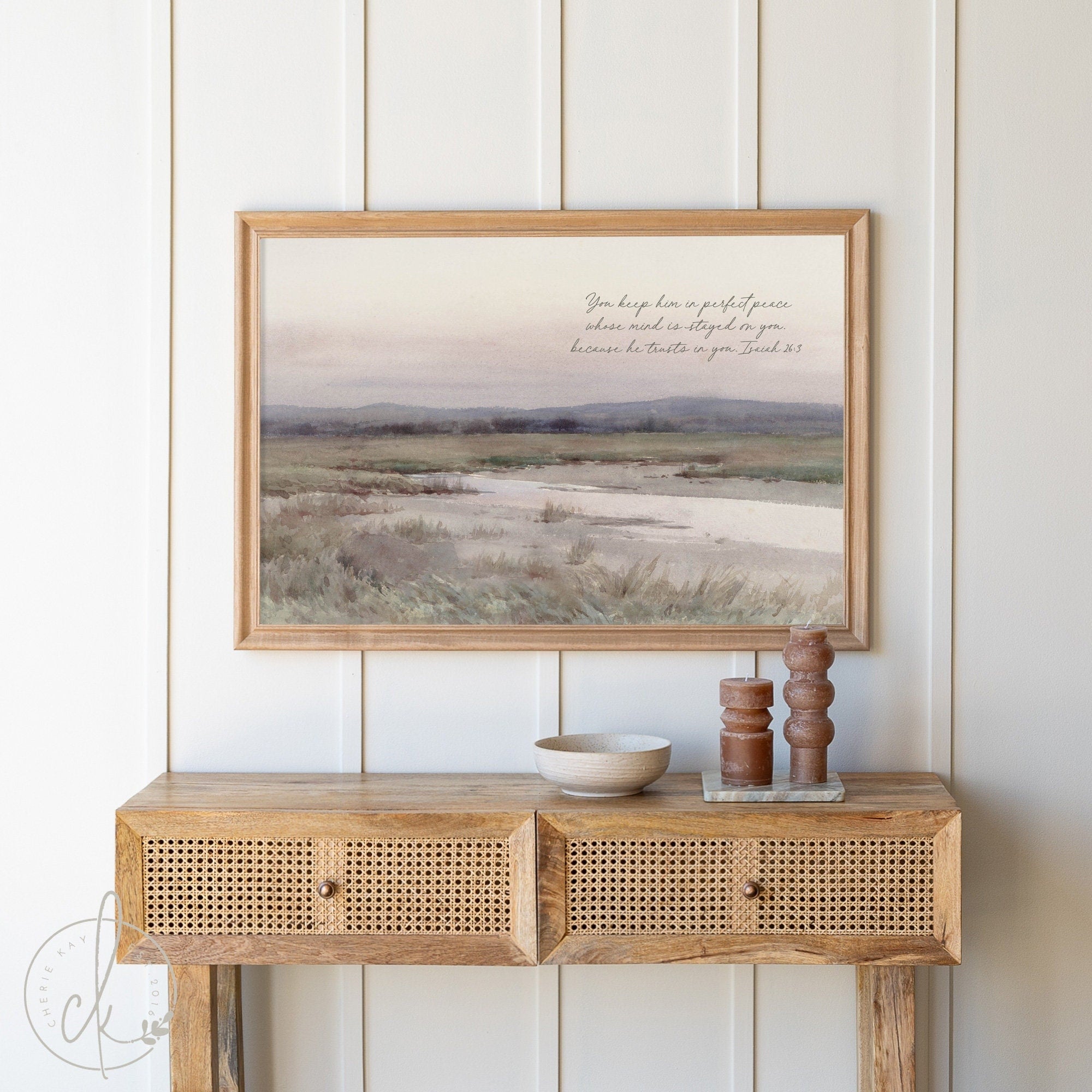 A wooden console table topped with a framed watercolor landscape art sign. The sign features Isaiah 26:3 in elegant script. The natural wood frame complements the farmhouse-style decor, with decorative items like a bowl and candlesticks on the table.