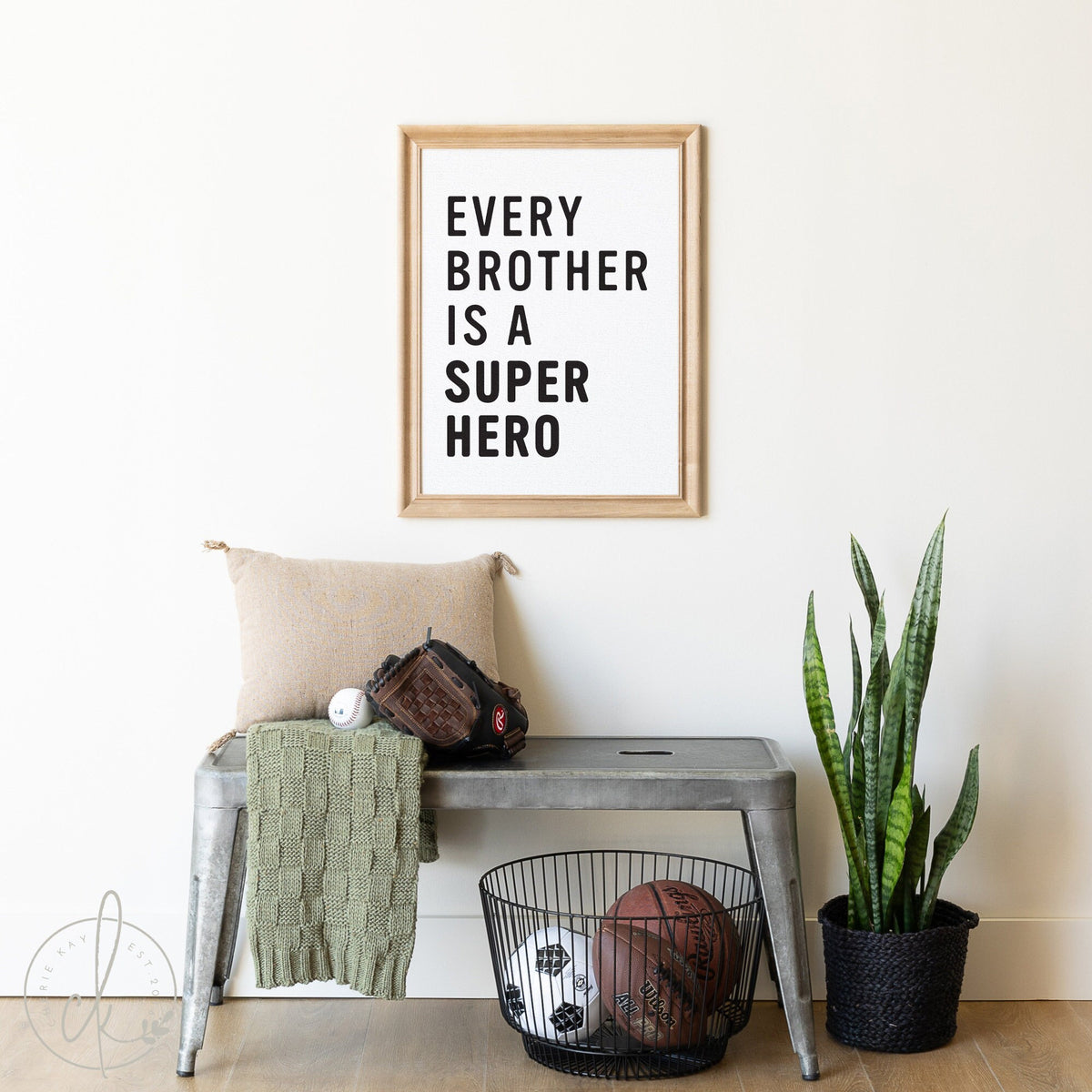 Framed sign with the text &amp;quot;Every Brother is a Super Hero&amp;quot; displayed above a gray metal bench. Below the bench, a wire basket holds a basketball and a soccer ball. A tall green plant in a black pot sits on the floor next to the bench