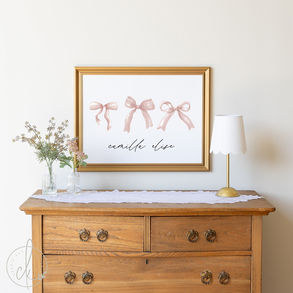 Elegant personalized nursery wall decor featuring three pink bows above the name &amp;#39;Camilla Elise&amp;#39; in cursive script, framed in a luxurious gold frame, styled on a wooden dresser with delicate floral accents.