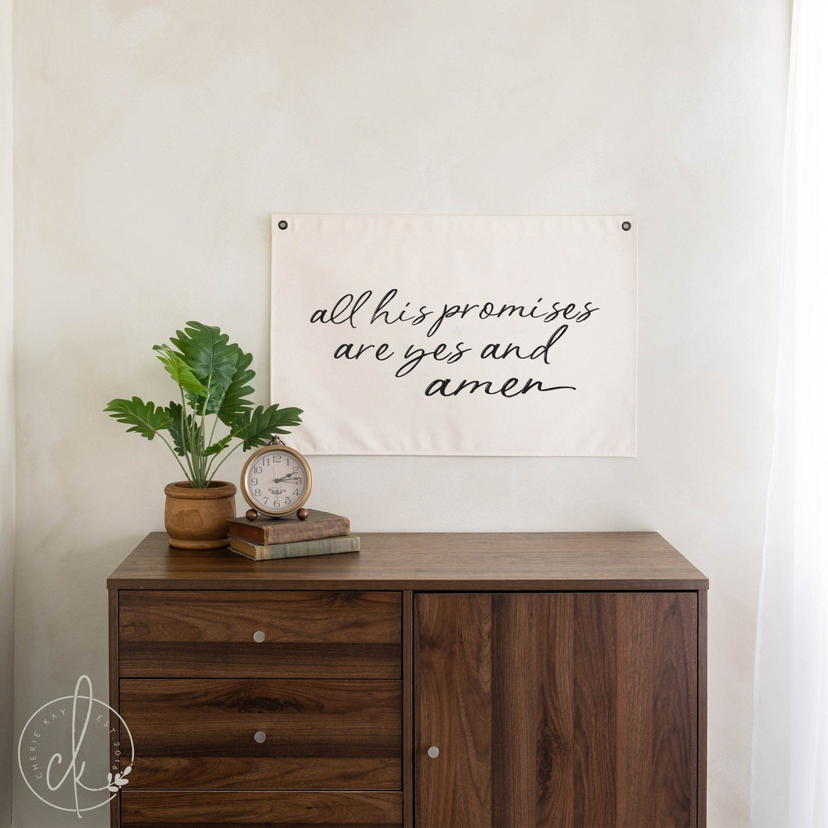 Decorative wooden dresser with a potted plant, vintage clock, and books, with a fabric wall banner hanging above it that reads All His Promises Are Yes and Amen in elegant script.