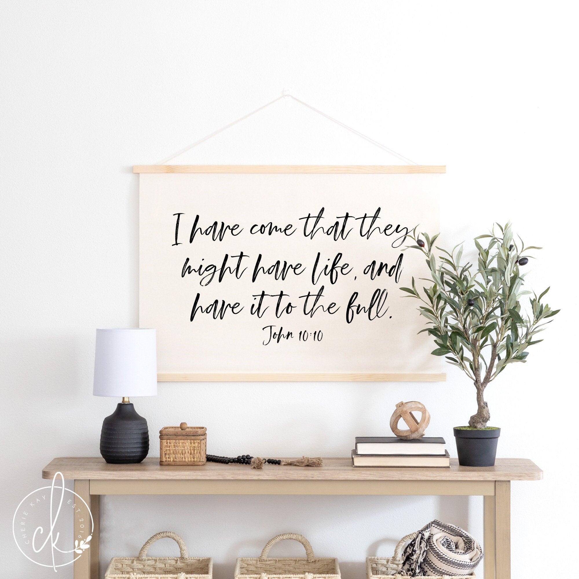 A modern living room featuring a canvas flag with the Bible verse John 10:10, I have come that they might have life, and have it to the full. The flag is framed with natural wood and hangs above a wooden console table decorated with a black lamp.