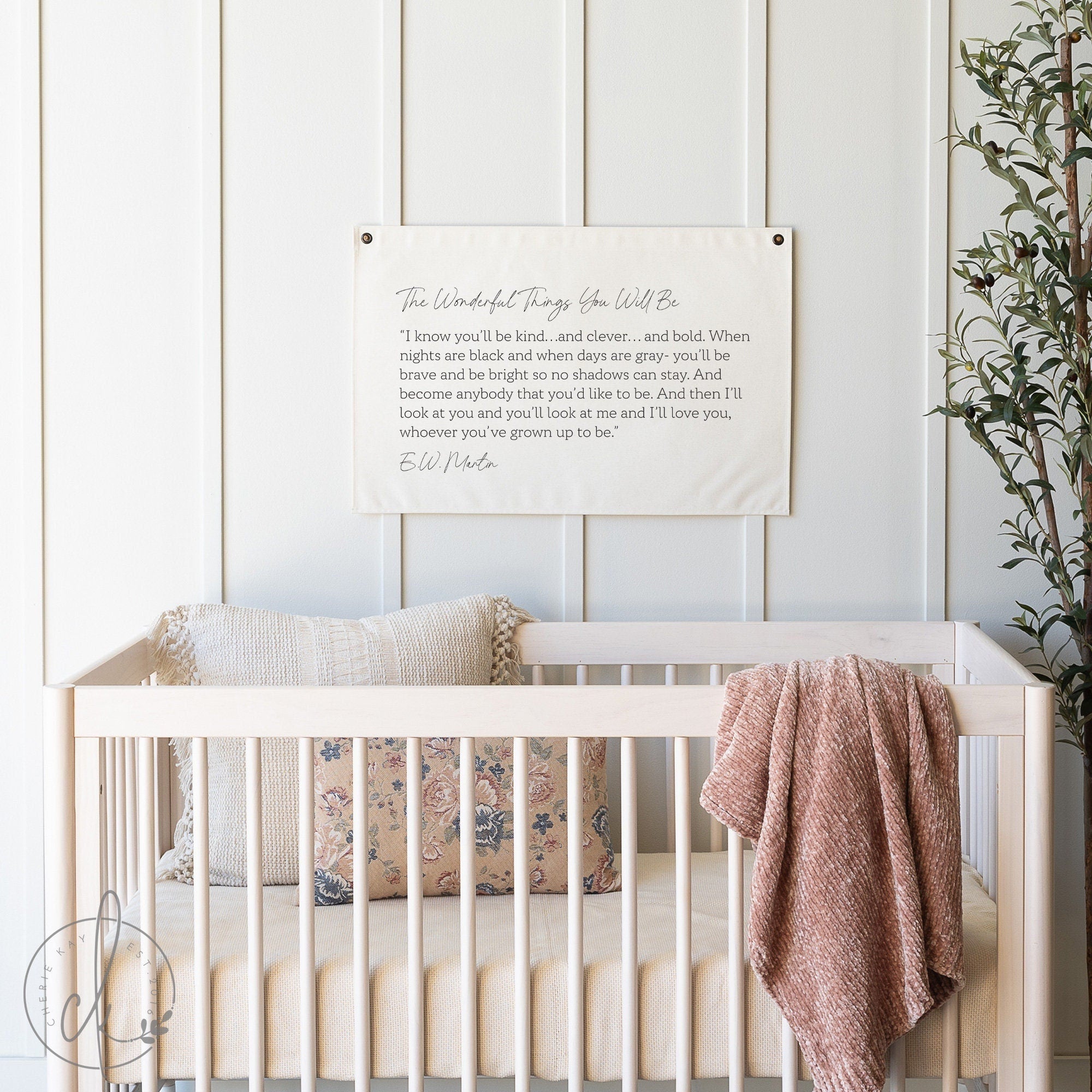 A cozy nursery features a white crib adorned with a floral pillow and a pink blanket draped over the side. Above the crib hangs a canvas flag with a quote by E.W. Martin printed in elegant script, adding an inspirational touch to the room&#39;s decor.