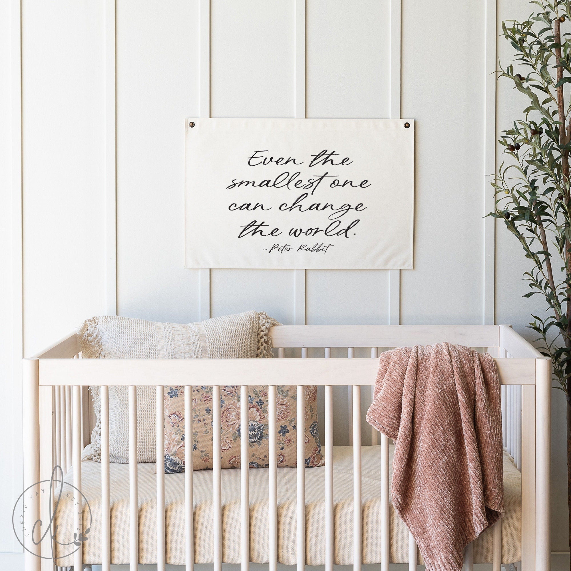 A cozy nursery with a wooden crib adorned with soft pillows and a pink knit blanket, featuring a wall hanging with the quote - Even the smallest one can change the world - by Peter Rabbit