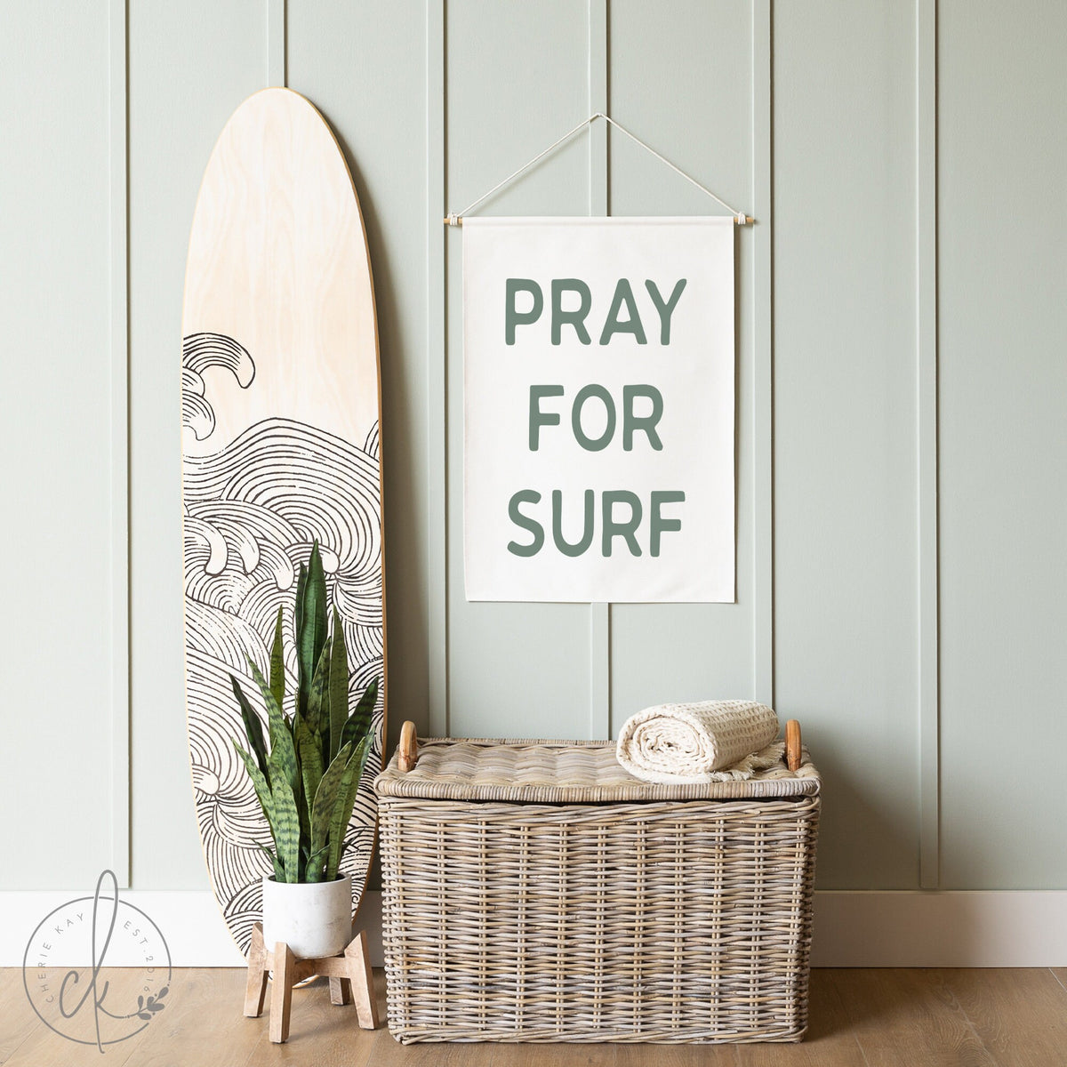 Pray for Surf canvas sign hanging on a light green paneled wall next to a surfboard and wicker basket, perfect for coastal decor, beach house styling, and surf-themed rooms. Minimalist ocean-inspired wall art for surf enthusiasts.