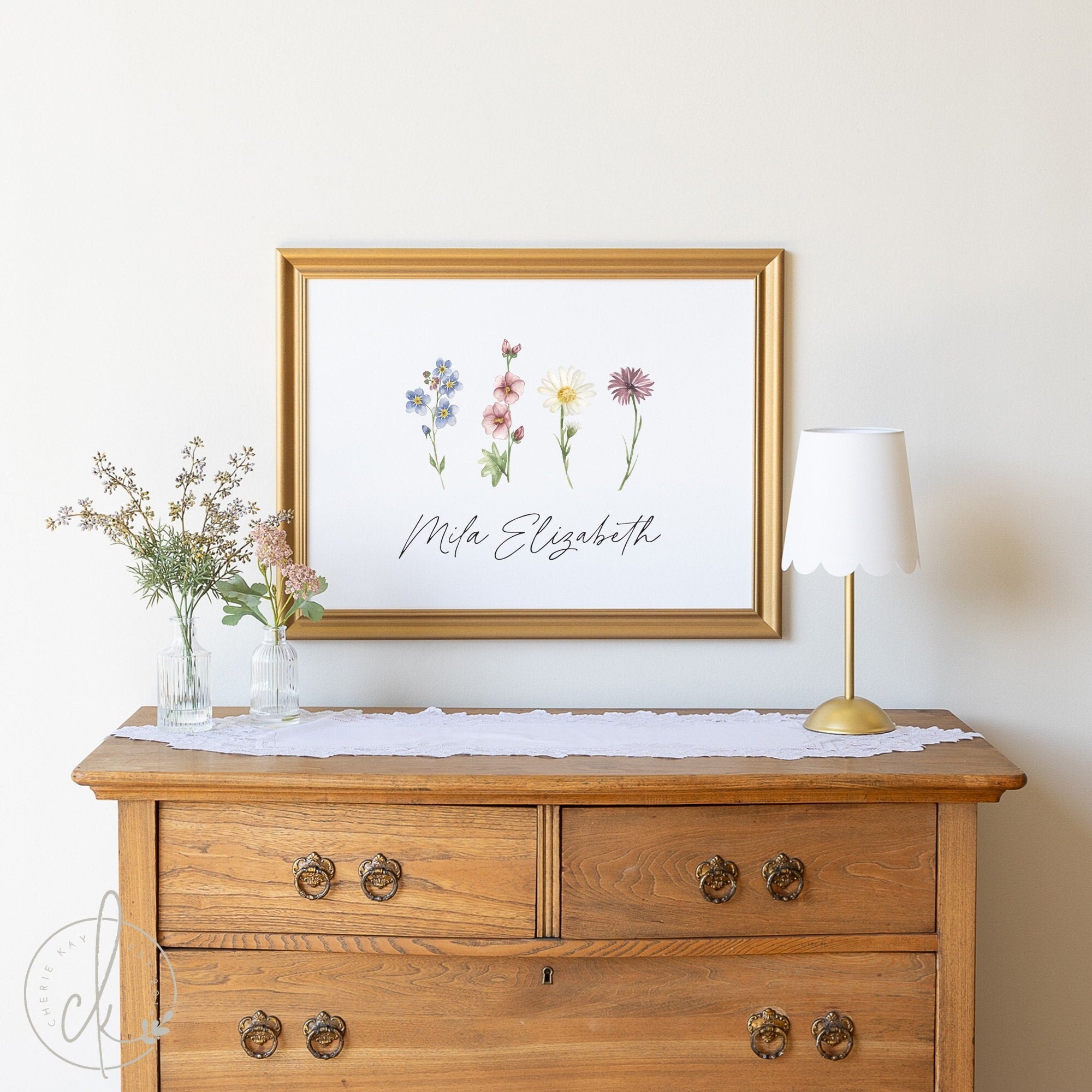 Gold-framed personalized girl name sign with wildflowers, perfect for nursery, bedroom, playroom, or teen room decor. Custom botanical floral art displayed above a wooden dresser with decorative items.