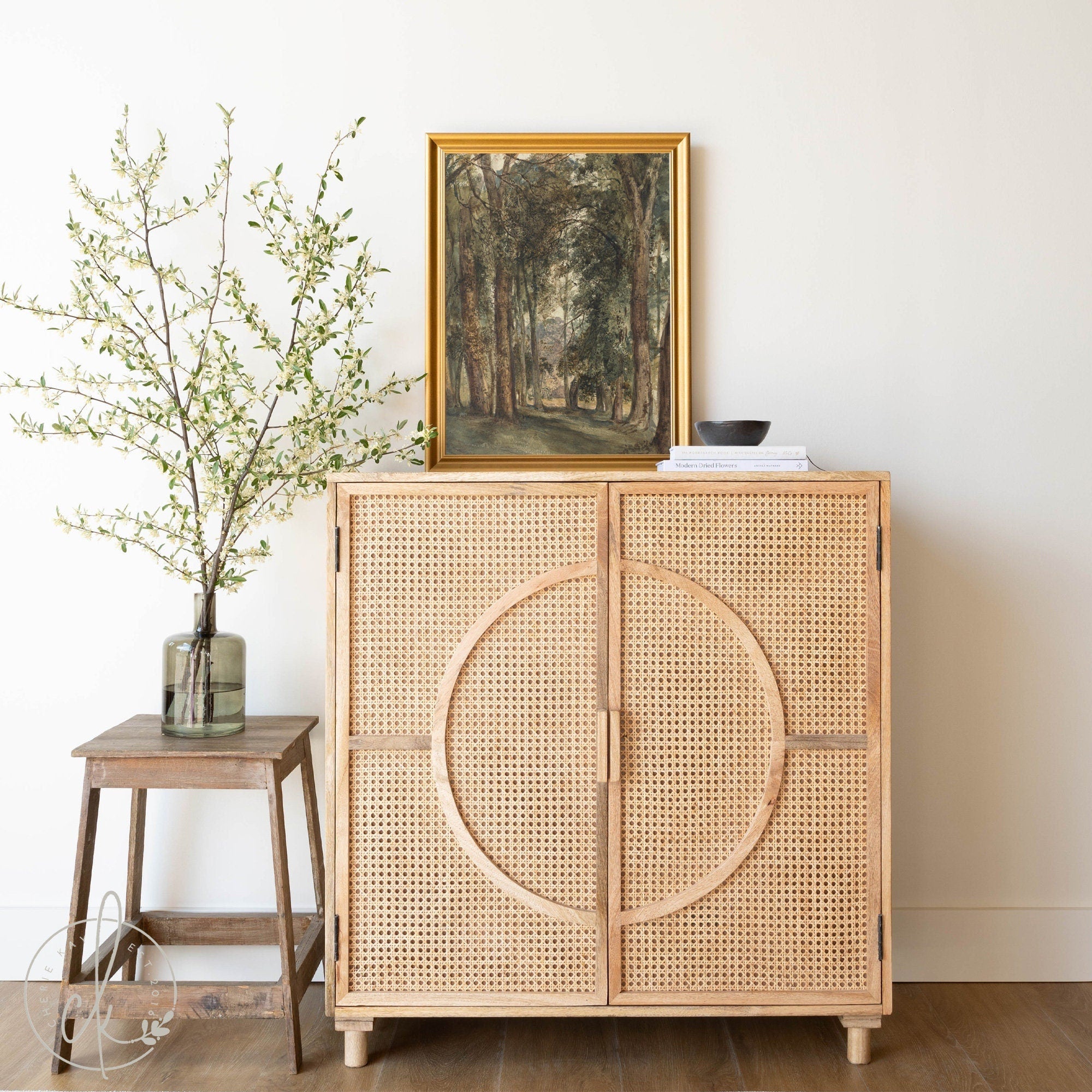 A beautifully styled living space featuring a vintage forest path landscape painting in a gold frame displayed on top of a natural rattan cabinet. Adding a touch of nature and elegance to the room decor. A blend of vintage and modern rustic elements.