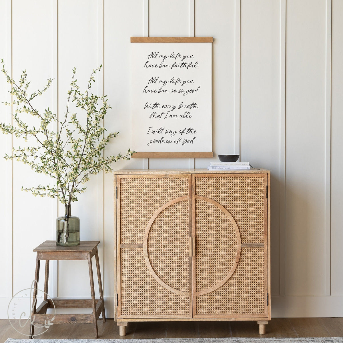 A serene home decor scene showcasing a tapestry with lyrics about faithfulness and goodness, hung above a rattan cabinet, beside a stool with a vase of budding branches.