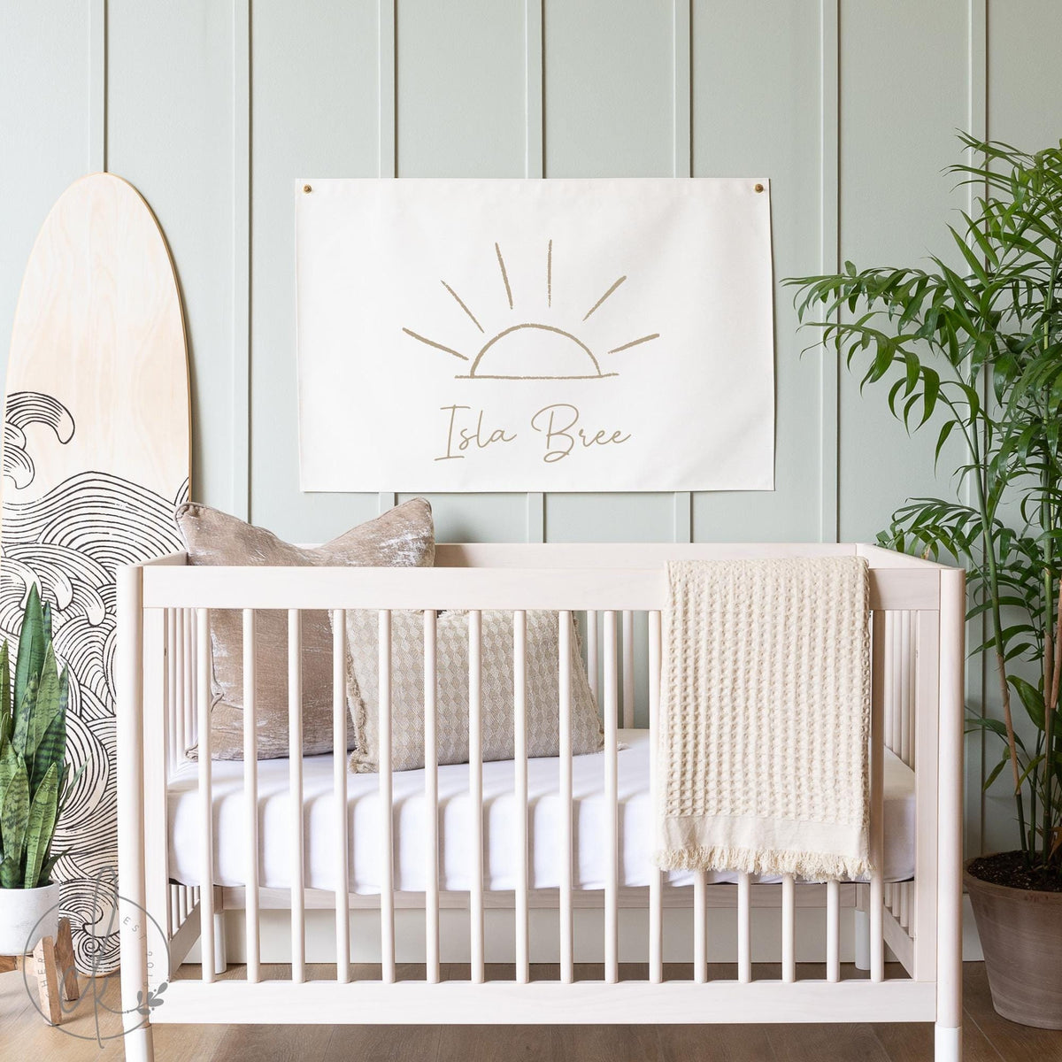 A stylish nursery featuring a light wooden crib with beige cushions and a cream blanket, set against a light green paneled wall. Above the crib hangs a personalized canvas flag with a minimalist sun sketch and the name &amp;quot;Isla Bree&amp;quot; written below.