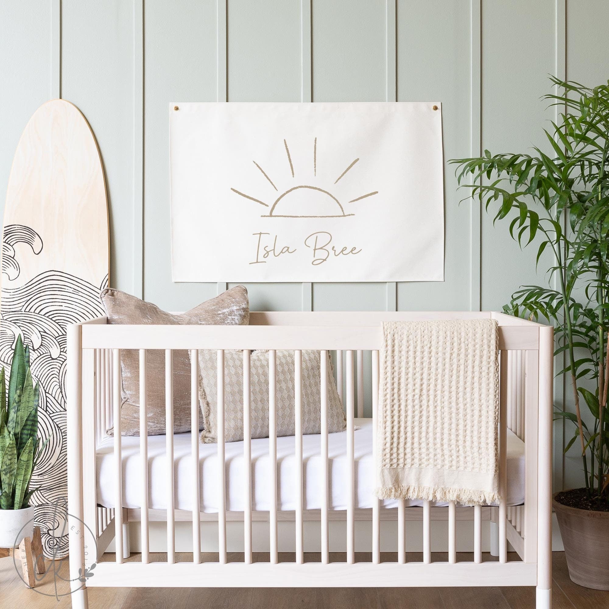 A stylish nursery featuring a light wooden crib with beige cushions and a cream blanket, set against a light green paneled wall. Above the crib hangs a personalized canvas flag with a minimalist sun sketch and the name &quot;Isla Bree&quot; written below.