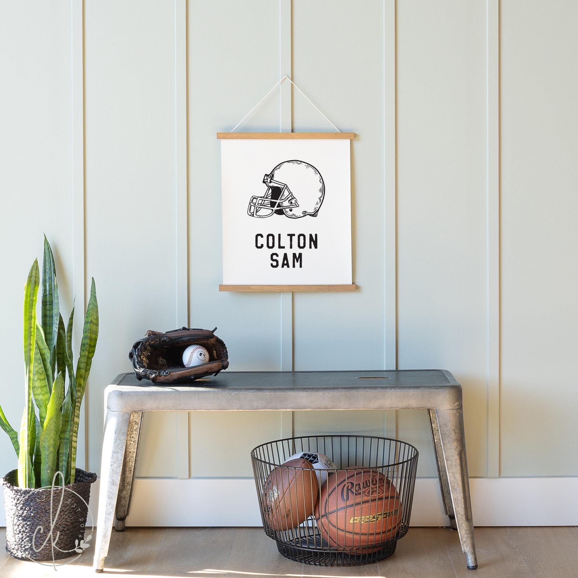 Personalized sports name sign featuring a black and white football helmet illustration with a custom name displayed underneath. The sign is hanging above a metal bench with a baseball glove and a wire basket containing balls.