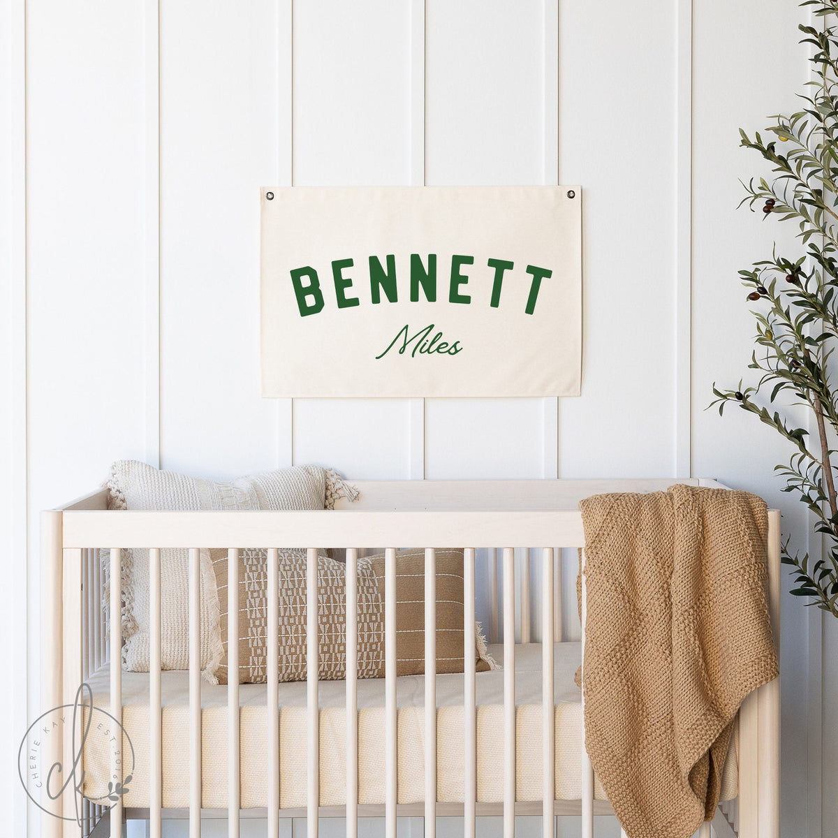 A personalized canvas name flag with the name Bennett Miles is displayed above a crib in a nursery room. The nursery features white walls, a crib with beige bedding, and a cozy blanket draped over the crib.