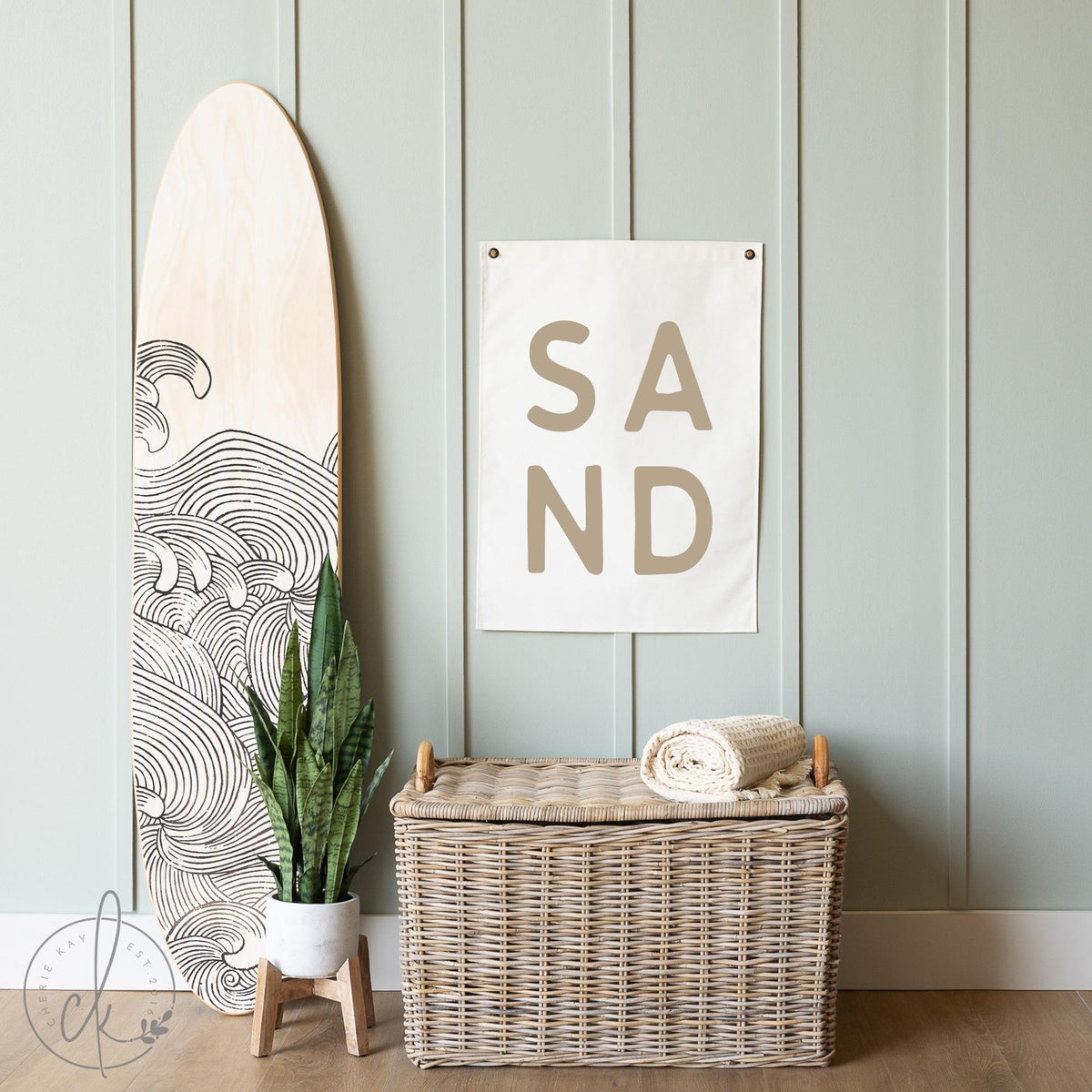 Coastal-themed room decor with a canvas sign featuring the word SAND in large capital letters, displayed on a light green paneled wall next to a surfboard. Ideal for beach house, coastal nursery, or ocean-inspired room decor.