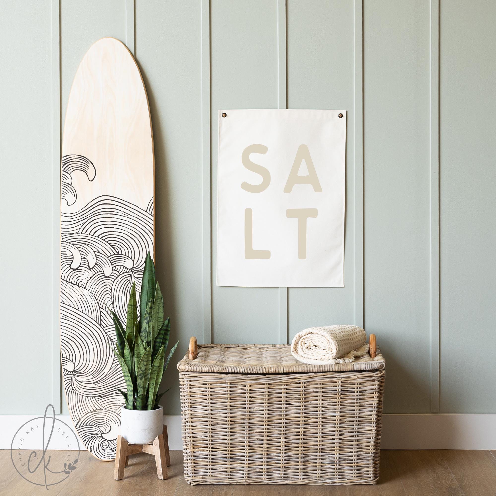 Coastal-themed room decor with a canvas sign featuring the word SALT in large capital letters, displayed on a light green paneled wall next to a surfboard. Ideal for beach houses, coastal nurseries, or ocean-inspired room decor.