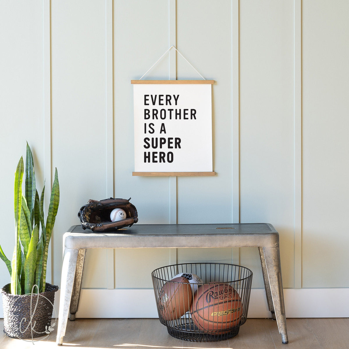 Canvas sign with the text &amp;quot;Every Brother is a Superhero&amp;quot; hanging on a light blue paneled wall. Below the sign is a metal bench with a baseball glove and ball on top, a wire basket with two basketballs underneath, and a potted snake plant on the side.