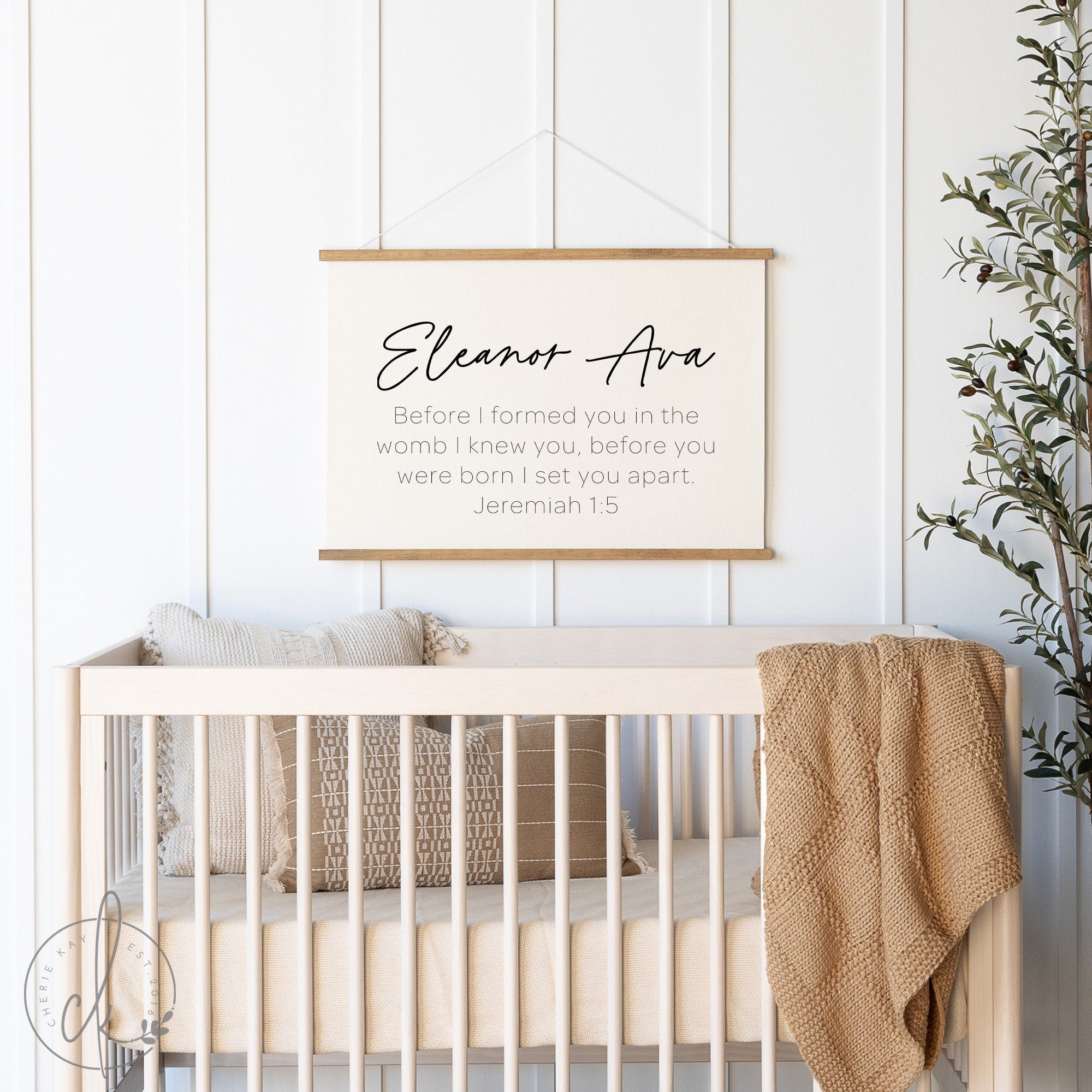 Personalized baby name sign hanging above a crib in a beautifully decorated nursery. The sign features the name Eleanor Ava and the verse Jeremiah 1:5. The nursery includes cozy pillows, a knitted blanket, and a potted plant.