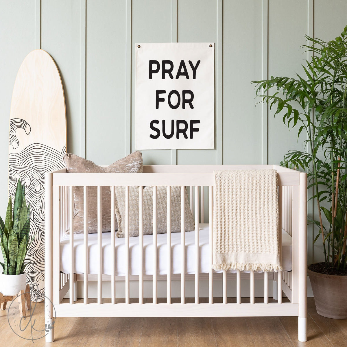 A coastal-themed nursery featuring a Pray for Surf flag hanging on a light green wall above a white crib. The decor includes a surfboard with wave designs, pillows, a cozy beige blanket, and various green plants, creating a beach-inspired ambiance.