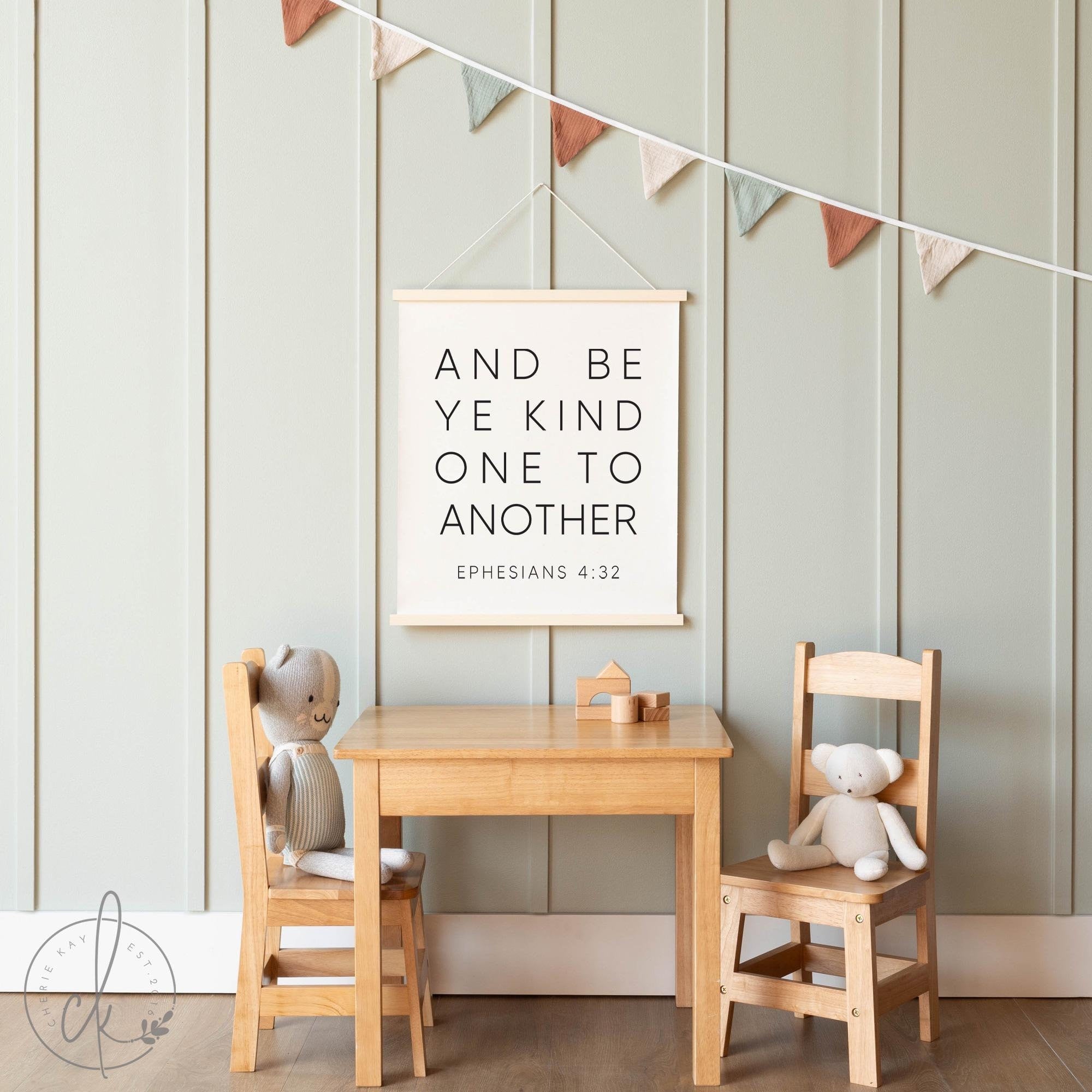 Childrens play area with a wooden table and chairs set against a soft green paneled wall. A banner with colorful fabric flags hangs above a framed sign displaying the Bible verse And be ye kind one to another, Ephesians 4:32.