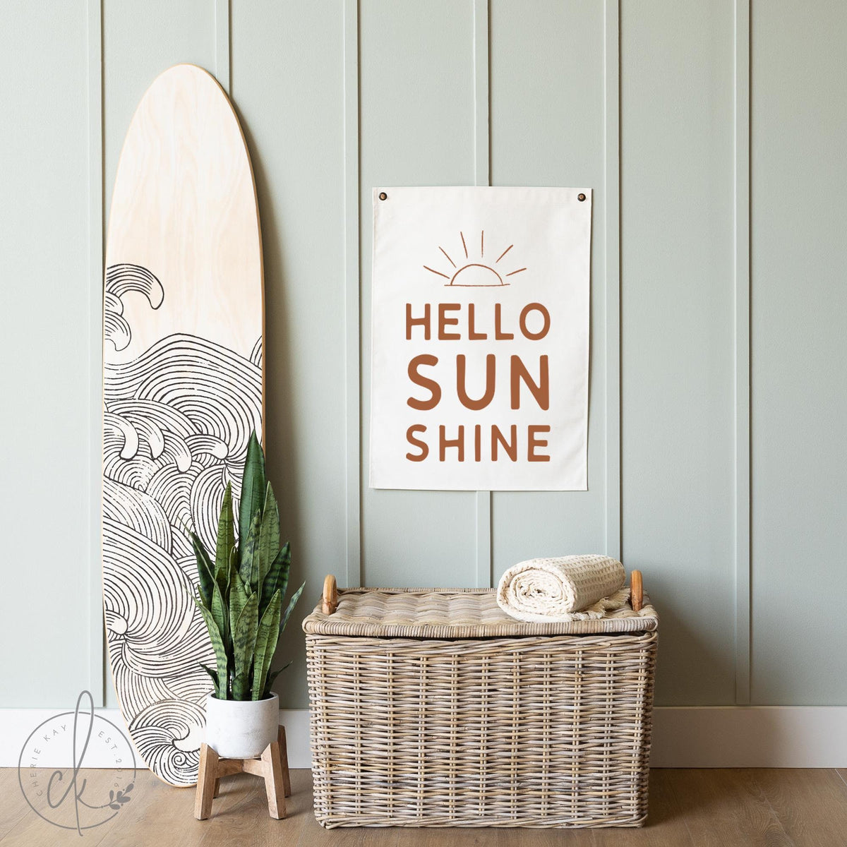 Hello Sunshine canvas flag with a sunrise sketch, perfect for coastal decor, beach houses, nurseries, and kids&amp;#39; rooms. Displayed next to a surfboard and wicker basket, creating a warm and inviting atmosphere.