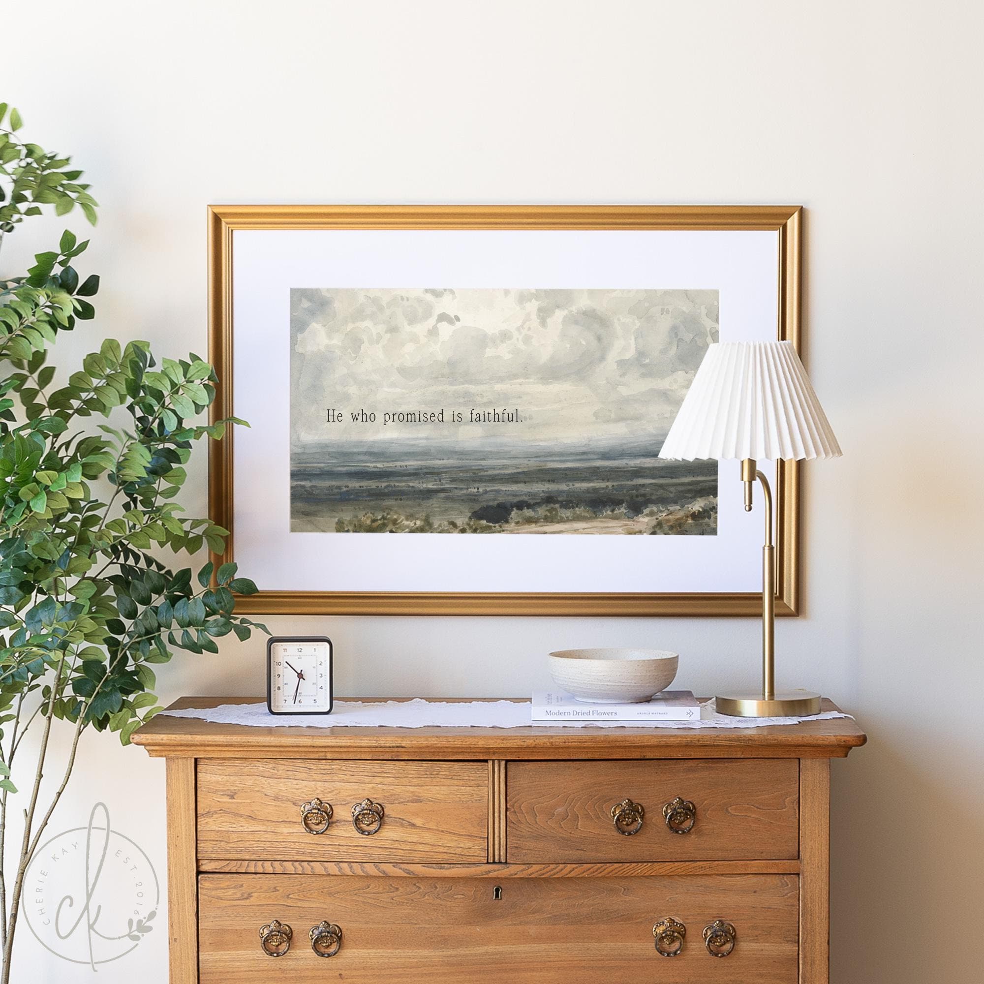 Framed vintage watercolor landscape art painting with the scripture. He who promised is faithful. Displayed above a wooden dresser, complemented by a lamp and potted plant.