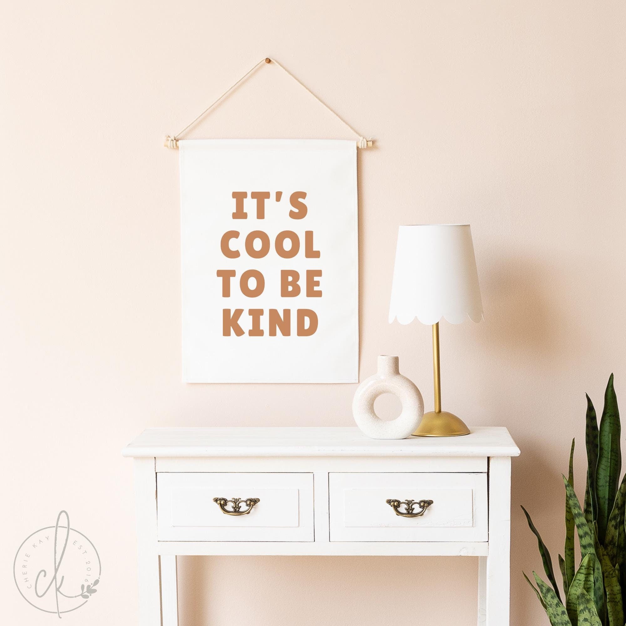 Canvas flag with It&#39;s Cool to Be Kind printed in bold letters, hanging on a neutral-toned wall above a white dresser, perfect for kids&#39; rooms, nurseries, and playrooms to inspire kindness and positivity.