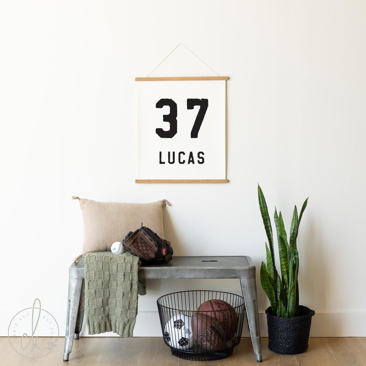 A personalized sports-themed sign featuring a large 37 and the name LUCAS displayed in bold, black typography on a neutral hanging sign. The sign is hung on a white wall above a metal bench with a baseball glove, a ball, and a green knit throw.