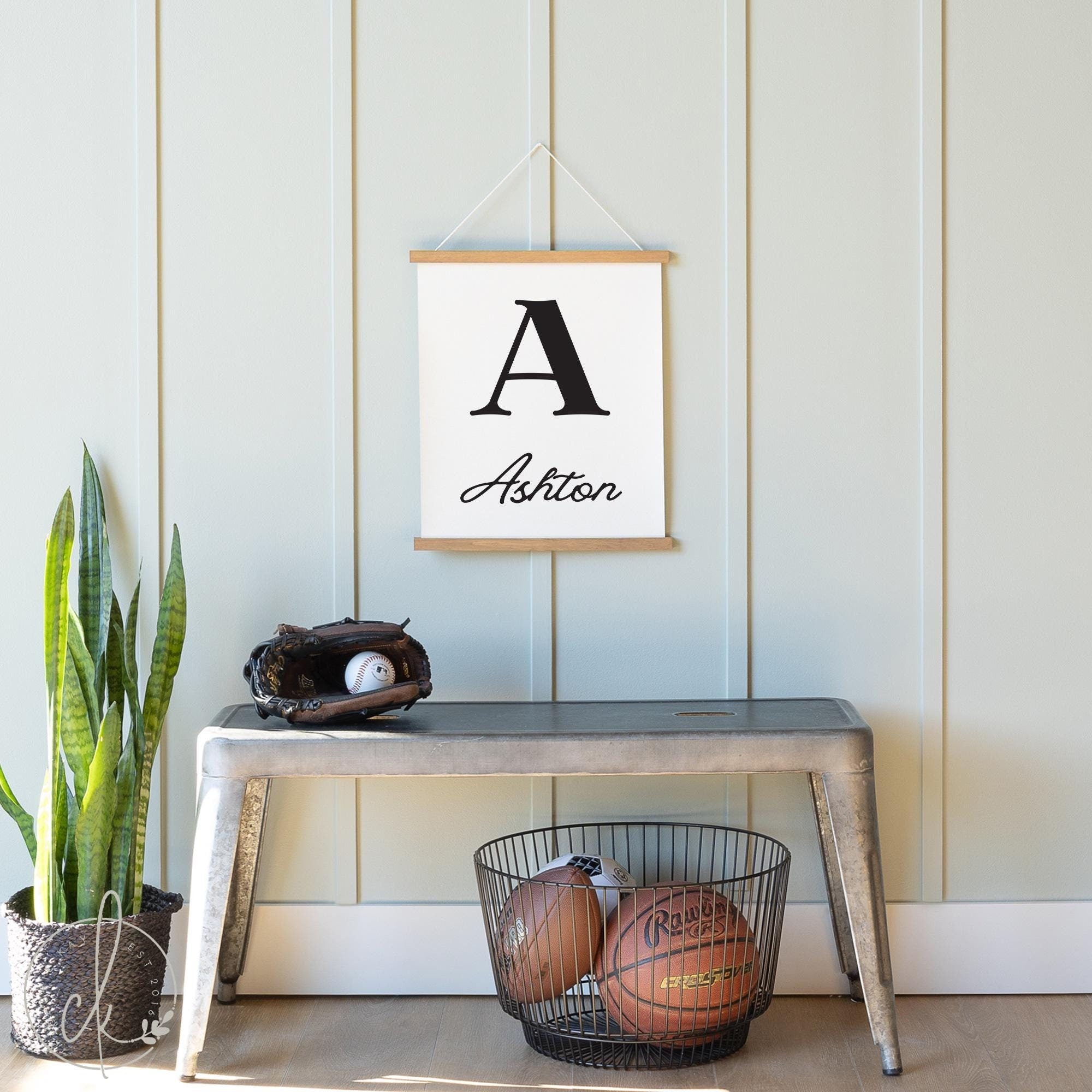 Personalized nursery canvas sign with bold initial and name hanging on a light-colored wall above a metal bench, surrounded by sports equipment and indoor plants, perfect custom baby room decor or gift for boys, or girls nurseries.