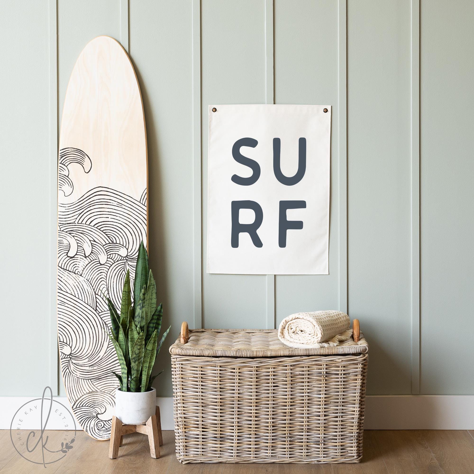 Coastal-themed room decor with a canvas sign featuring the word SURF in large capital letters, displayed on a light green paneled wall next to a surfboard. Perfect for beach house, coastal nursery, or ocean-inspired room decor.