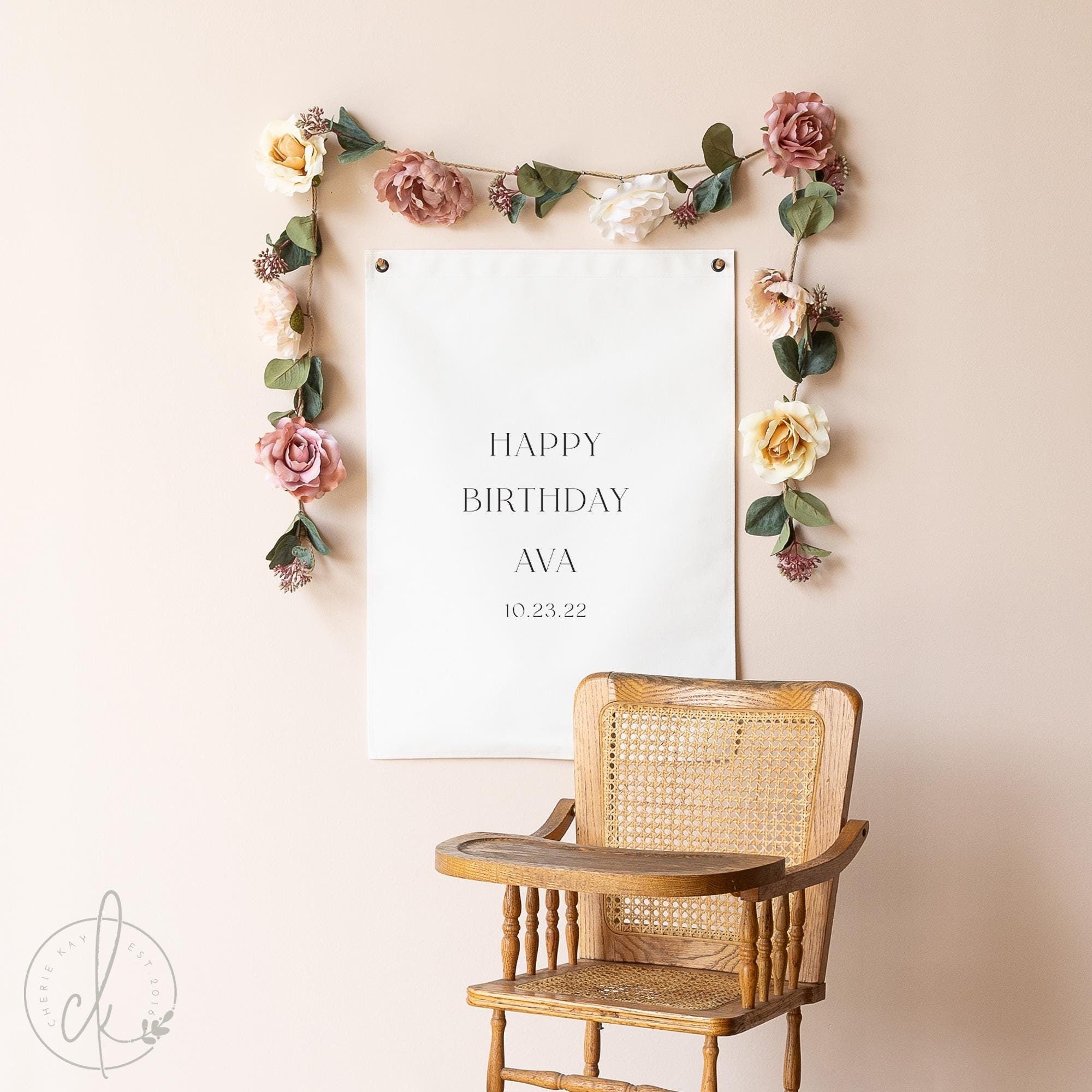 A personalized birthday banner hanging on a wall, displaying Happy Birthday with the custom date. The banner is surrounded by a decorative floral garland, and a vintage wooden high chair is positioned in front of it.