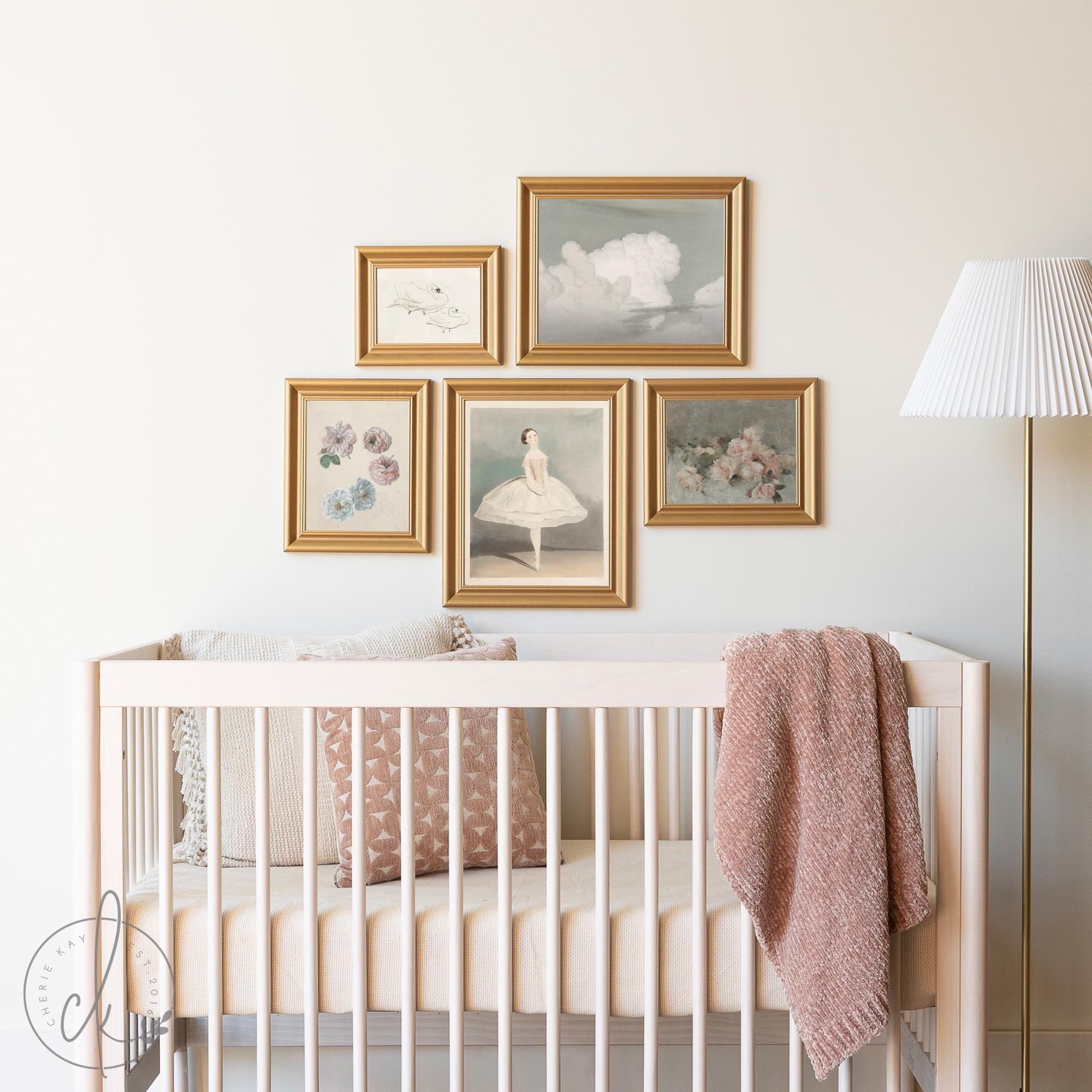 A nursery with a light wooden crib, soft bedding, and a pink blanket. Above the crib, a gallery wall features five gold-framed art prints, including florals, a ballet dancer, and clouds. A white floor lamp stands nearby.