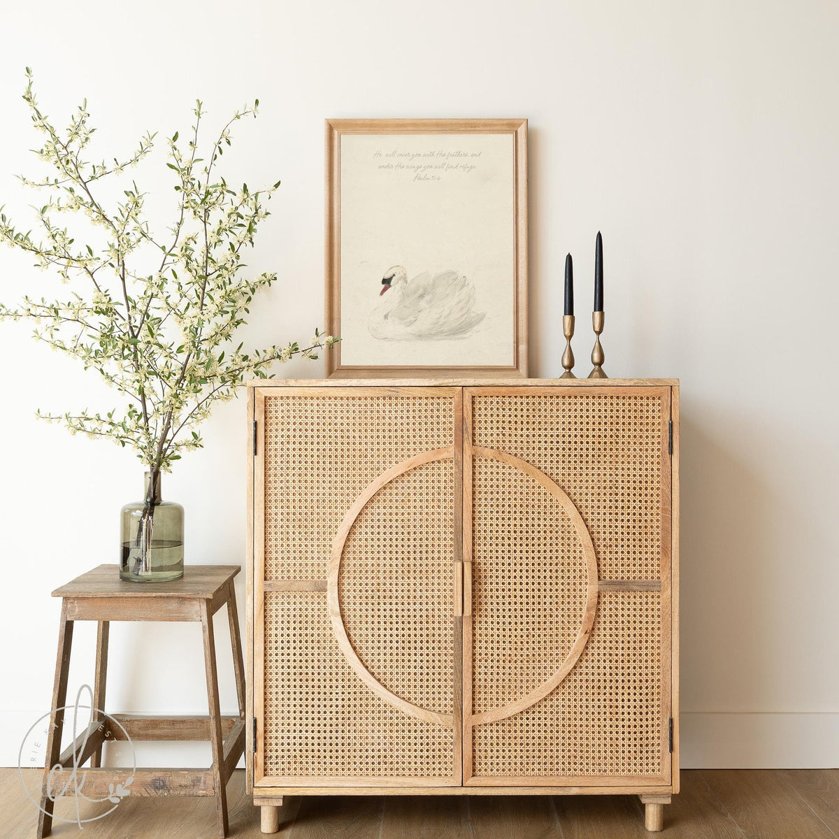 Minimalist decor with a framed swan illustration on a rattan cabinet, flanked by black candles and a glass vase with leafy branches, against a white wall.