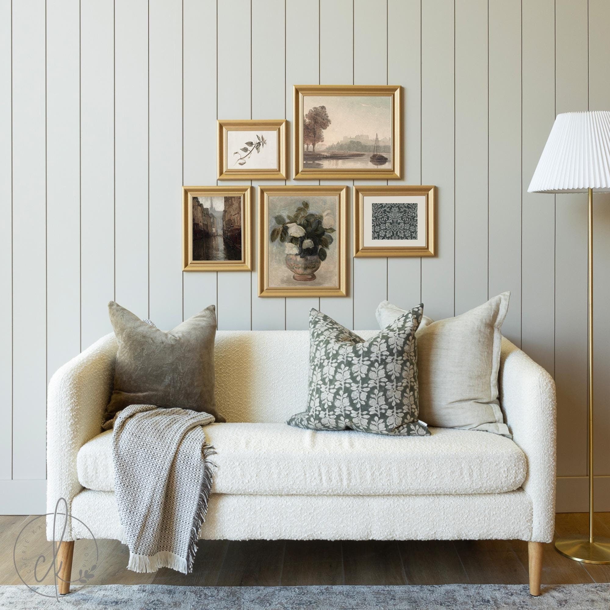 Curated vintage gallery wall featuring framed botanical and landscape art prints in gold frames, styled above a cozy cream sofa with decorative pillows in a living room setting.