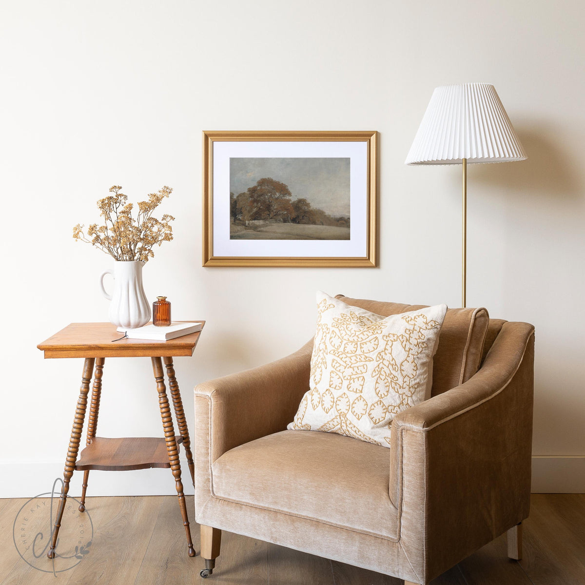 Cozy living room corner with framed fall landscape art, beige armchair, wooden side table, and dried flowers.