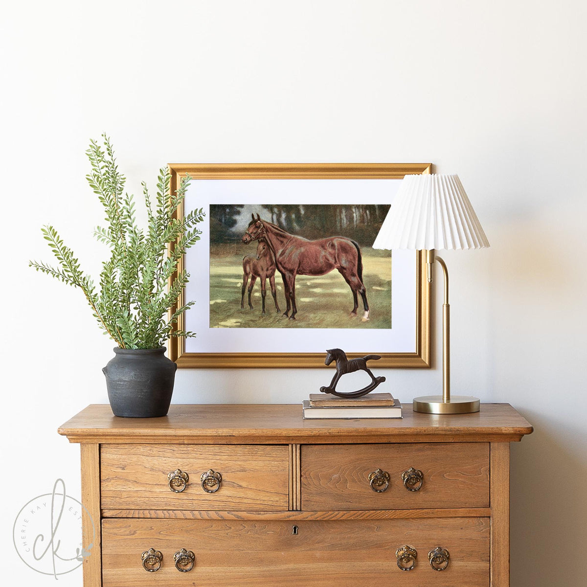 A wooden dresser topped with a gold-framed painting of a horse and foal. A black vase with greenery and a small white-shaded lamp sit beside a decorative horse figure. The scene has a warm and classic feel.