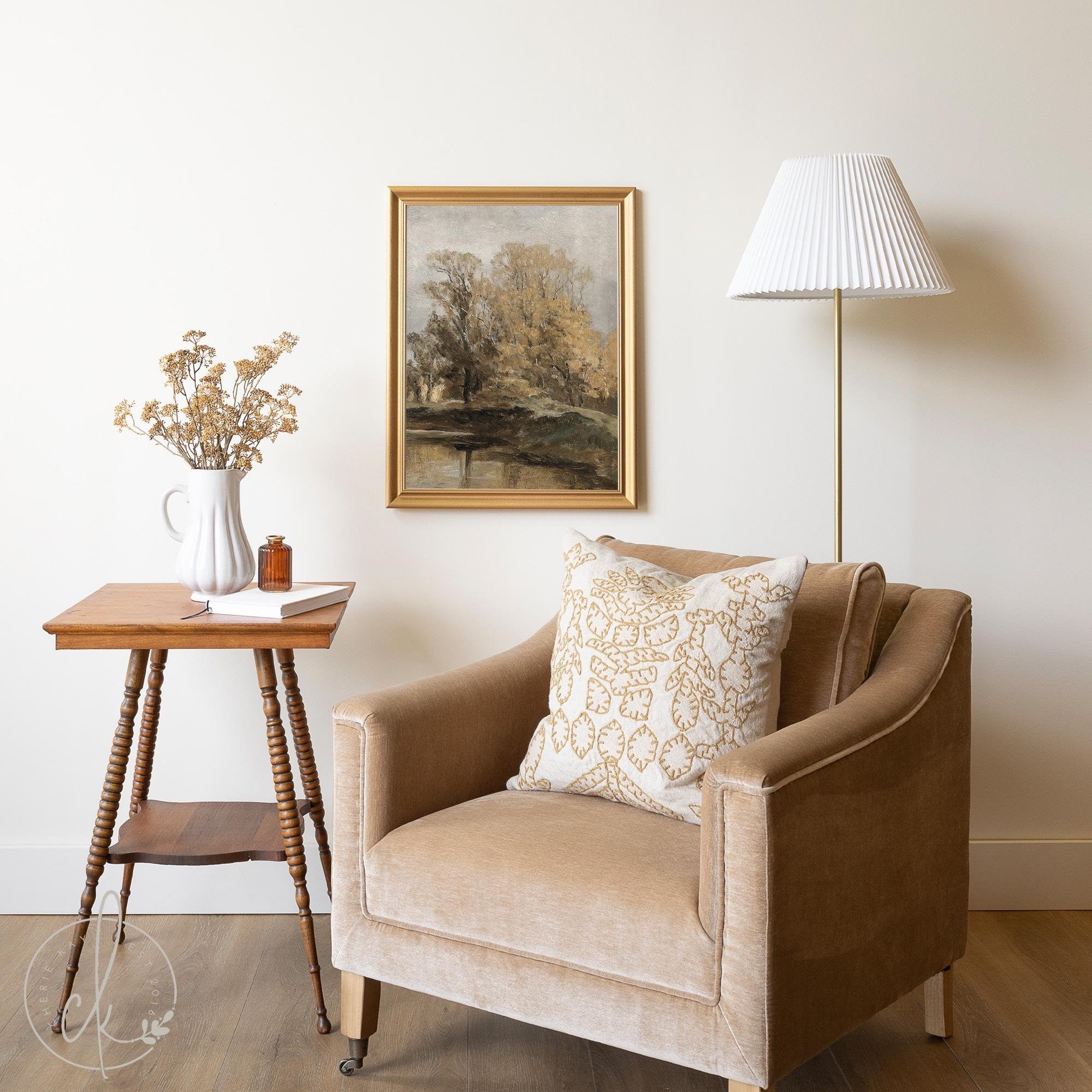 A cozy living space featuring a beige armchair with a patterned cushion, a small wooden side table with dried flowers in a white pitcher, and a gold-framed landscape painting on the wall. A floor lamp with a pleated shade adds warmth to the room.