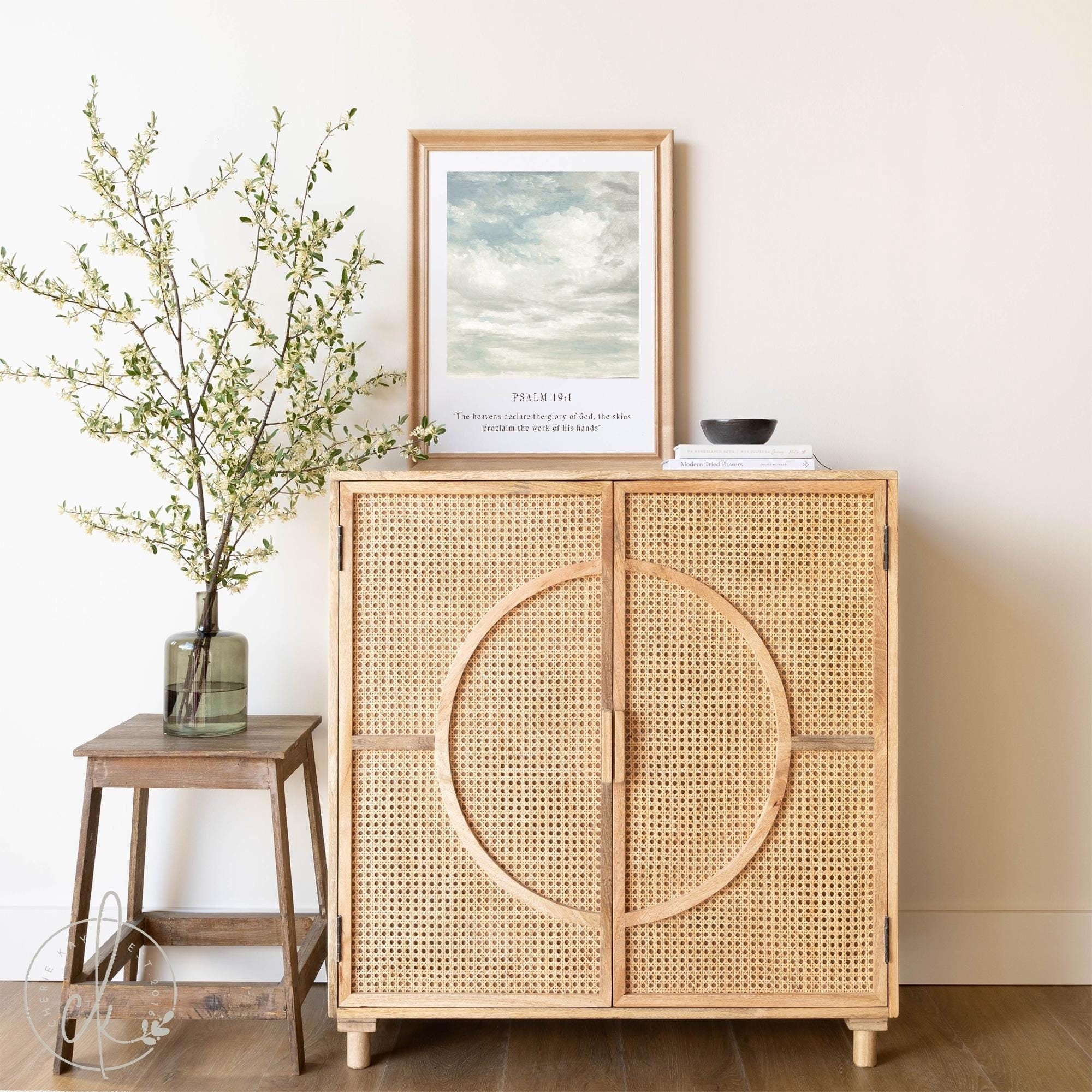 Wooden cabinet with framed Psalm 19:1 cloud illustration, a vase with branches on a stool, and minimal decor in a serene, natural setting.