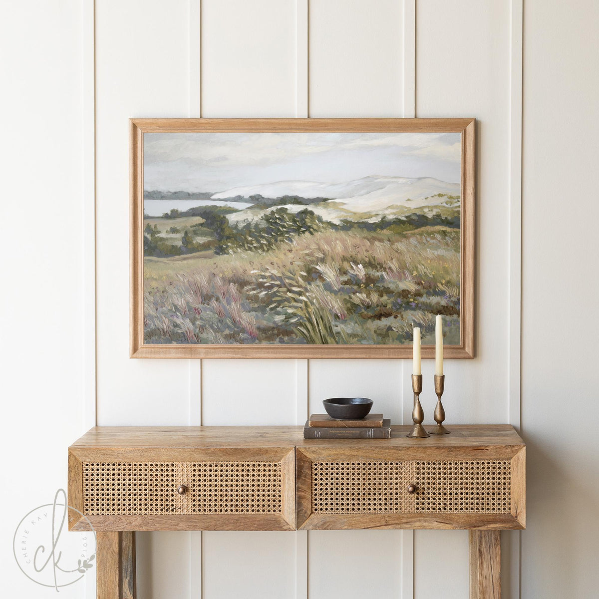 Framed landscape painting featuring rolling hills and greenery, displayed above a rustic wooden console table with rattan drawers, styled with two brass candlesticks and a small bowl in a cozy, neutral-toned room.