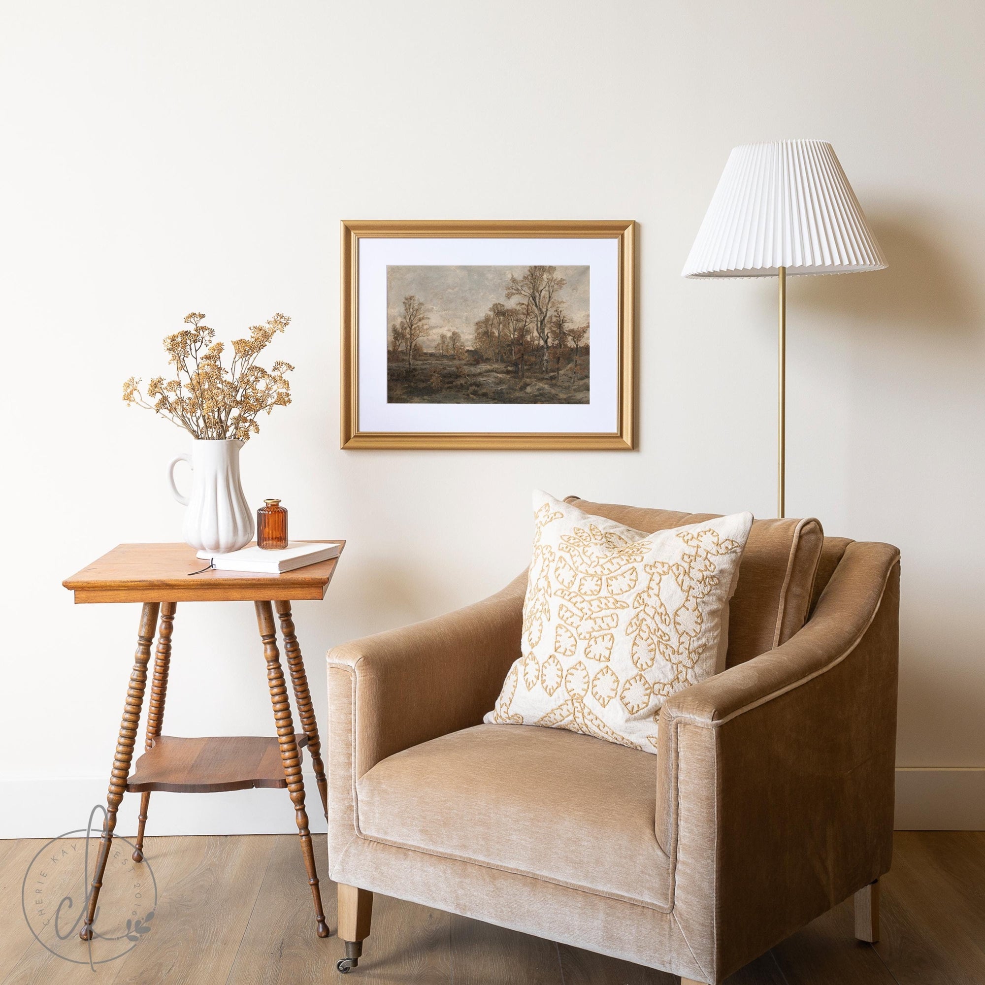 Living room with framed vintage landscape art in a gold frame, beige armchair, and wooden side table with dried flowers.