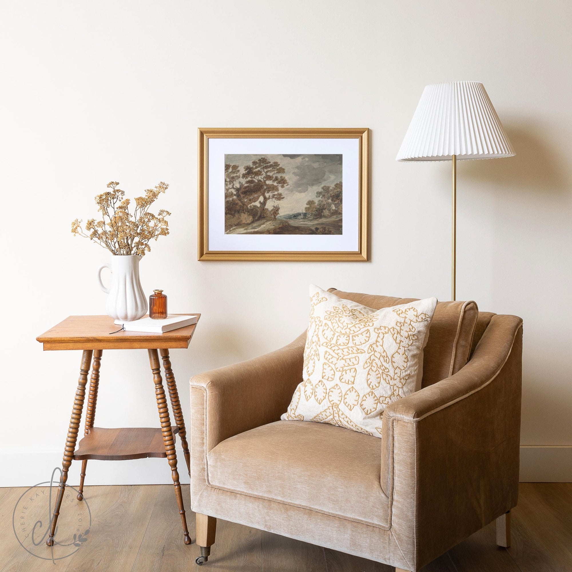 Framed fall landscape art in a gold frame above a cozy armchair with a cushion, side table, and floor lamp in a warm, inviting setting.