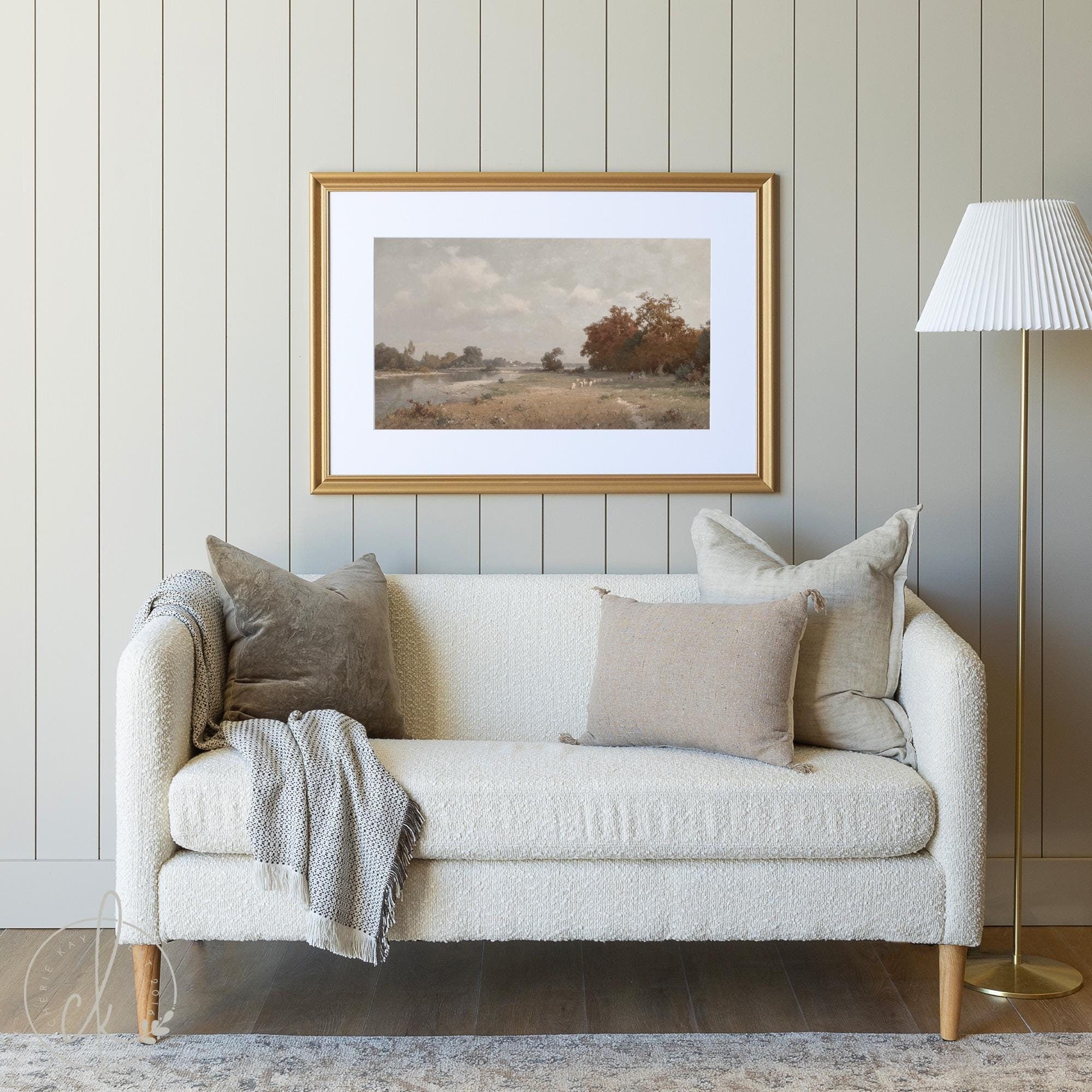 A cozy living room featuring a light beige sofa with soft pillows and a gray throw. A landscape painting of a serene autumn scene hangs on a light gray paneled wall. A white-shaded lamp stands on the right, adding to the minimalist, calm ambiance.