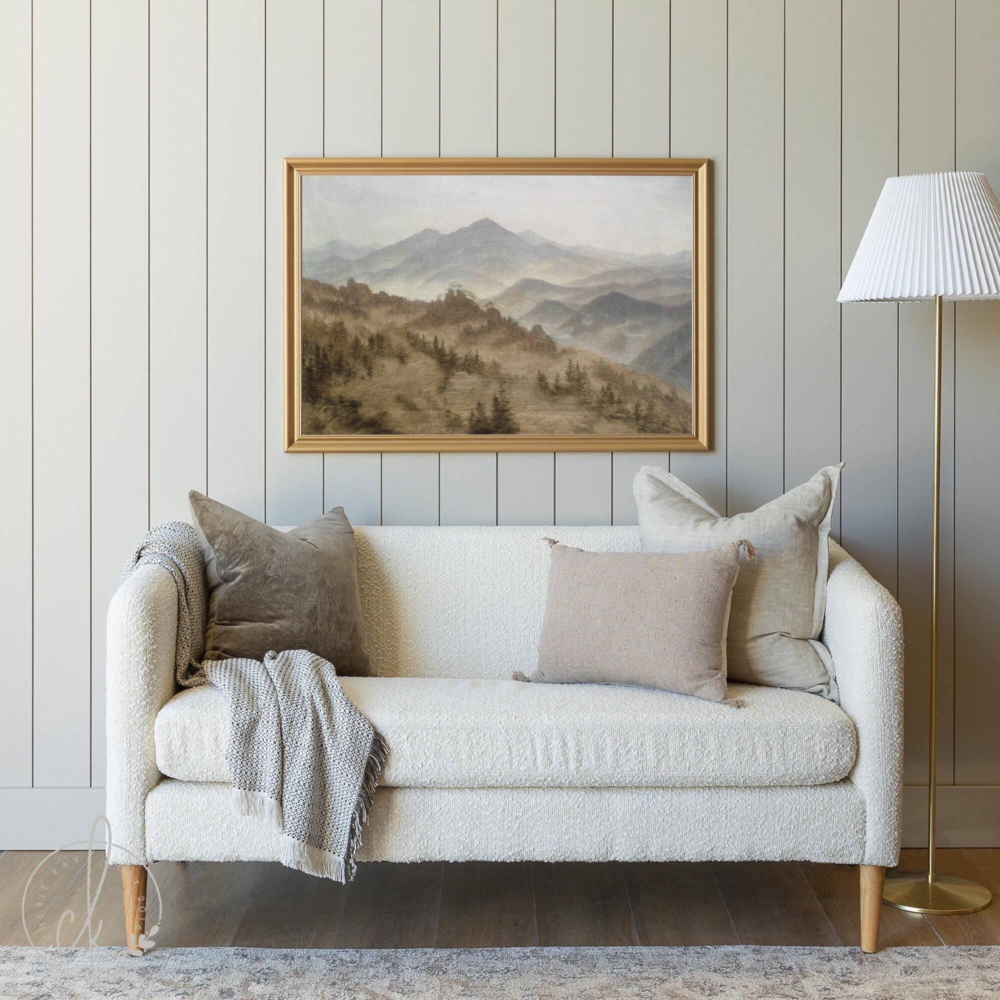 A cozy living room with a light beige sofa, soft pillows, and a gray throw. A gold-framed mountain landscape painting hangs on a light gray paneled wall, complemented by a white-shaded lamp on the right. The room has a calm and minimalist style.