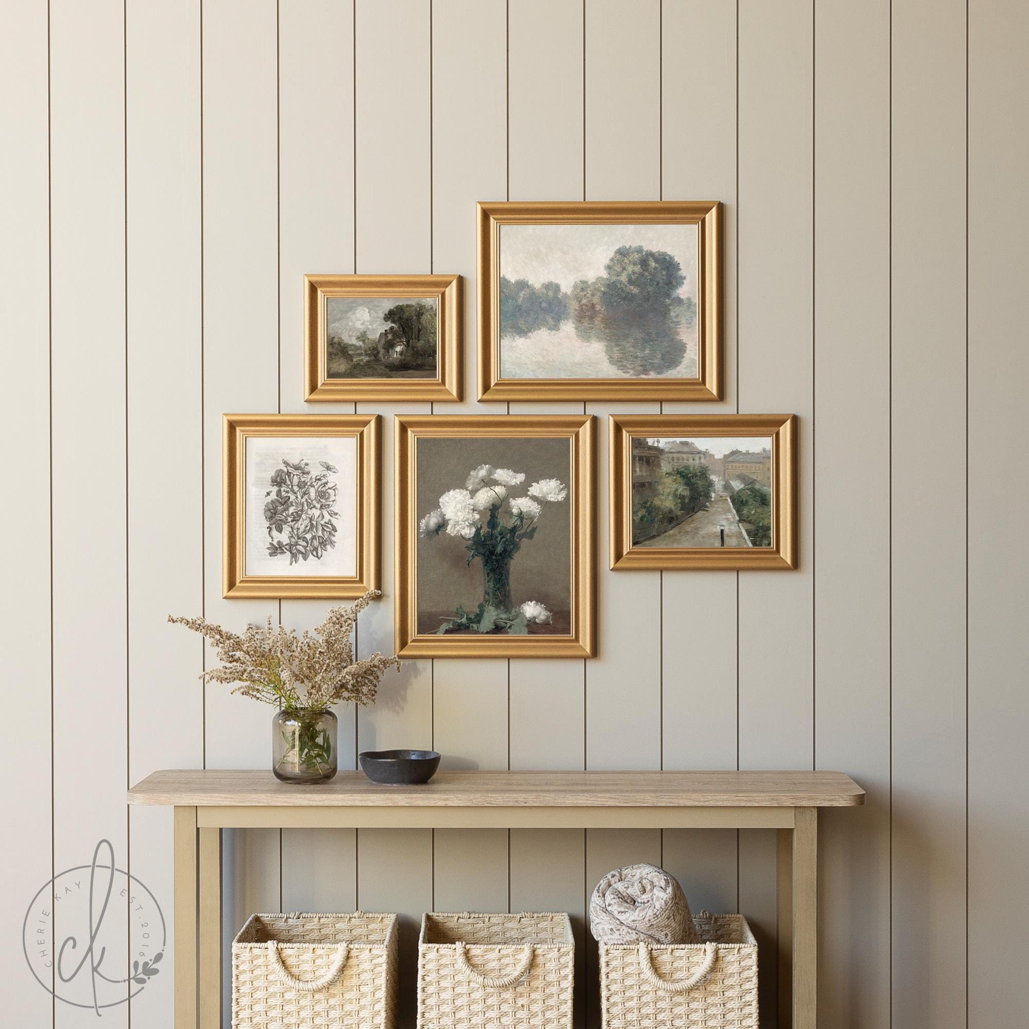 Gallery wall featuring five gold-framed art pieces, including vintage botanical sketches, serene landscapes, and floral prints, displayed on a light beige shiplap wall above a rustic wooden console with woven storage baskets and decorative accents.