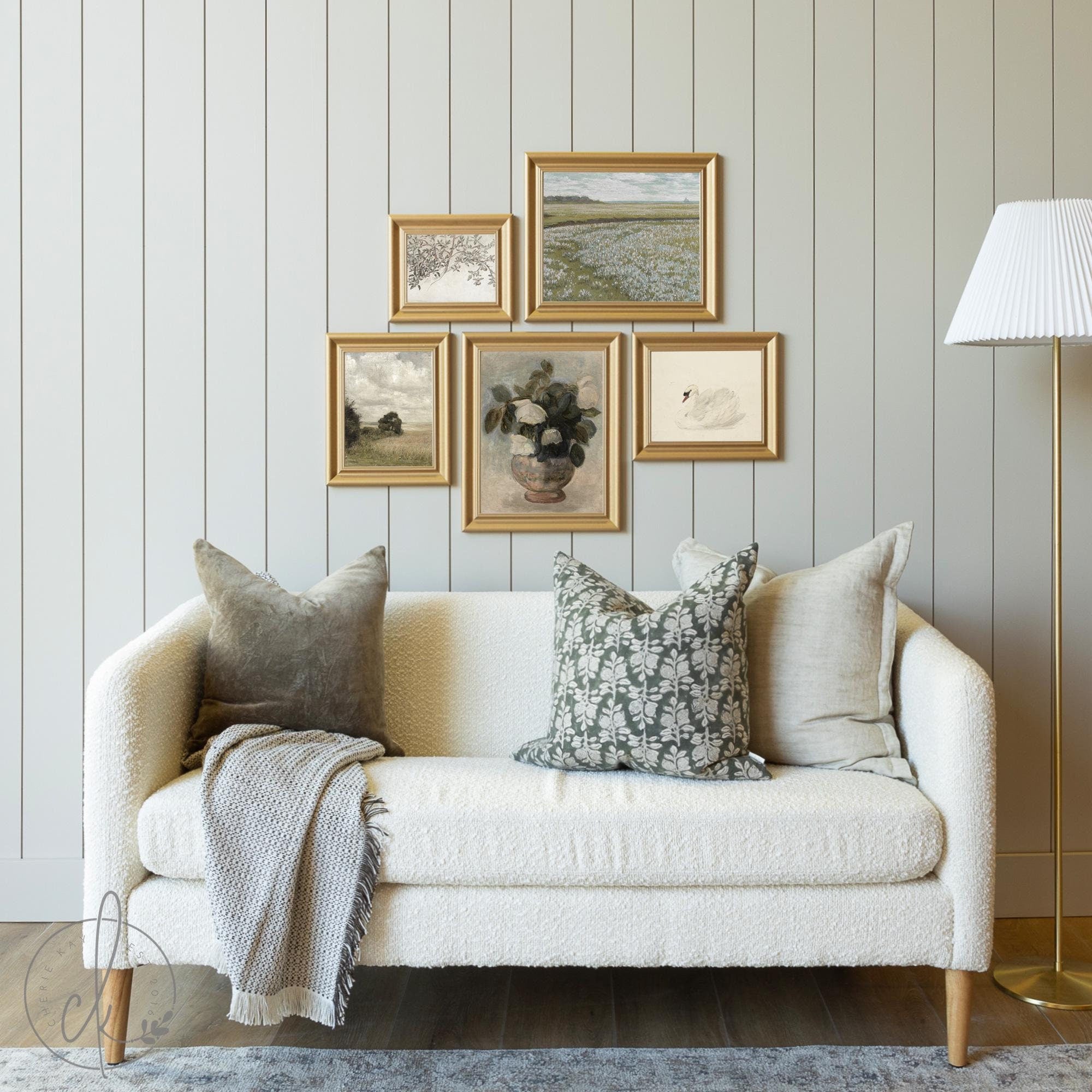 Cozy living room with a rustic gallery wall featuring five gold-framed art prints, including vintage landscapes, botanicals, and classic artwork, above a neutral sofa with accent pillows and a throw blanket.