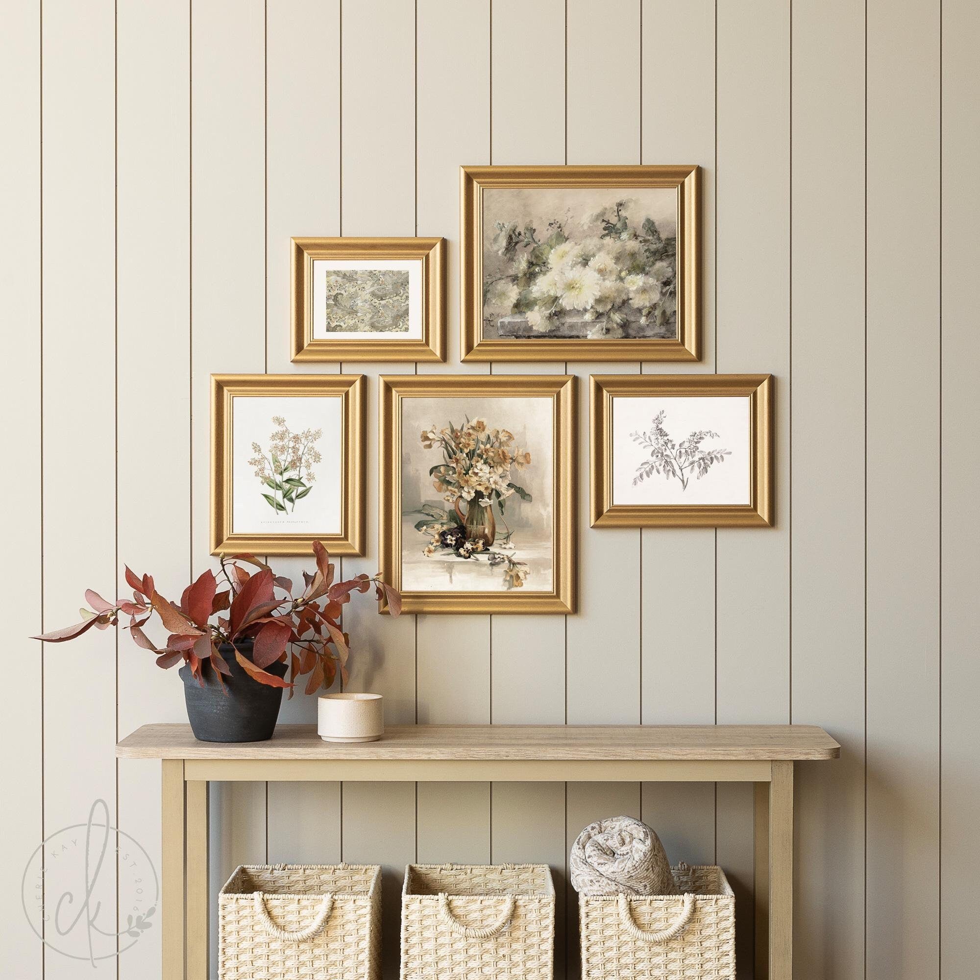 Vintage-inspired gallery wall with five framed floral and botanical prints in gold frames, displayed on a light wood paneled wall above a minimalist console table with woven storage baskets.