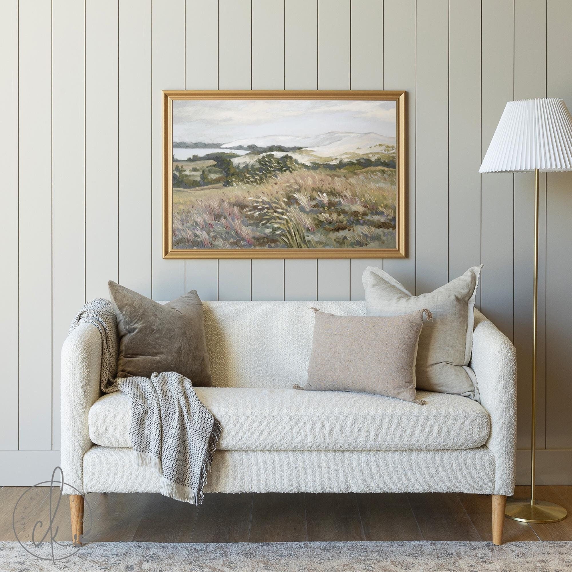 A cozy living room with a beige textured couch, neutral pillows, and a gray knit throw. A gold-framed landscape painting hangs on a light gray paneled wall. A white-shaded floor lamp stands to the right, creating a serene and minimalist space.