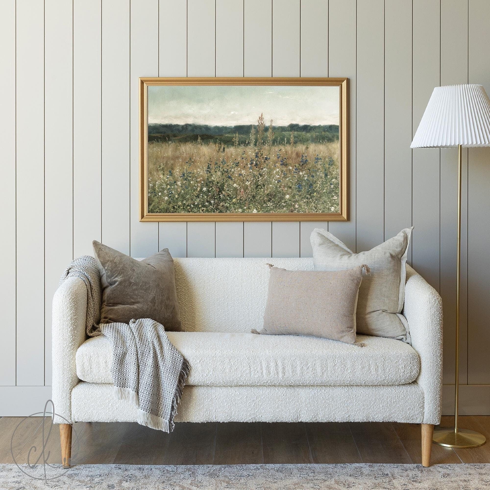 A cozy living room with a cream sofa, neutral pillows, a gray blanket, and a framed meadow painting on a muted paneled wall. A gold floor lamp with a white shade stands to the side.