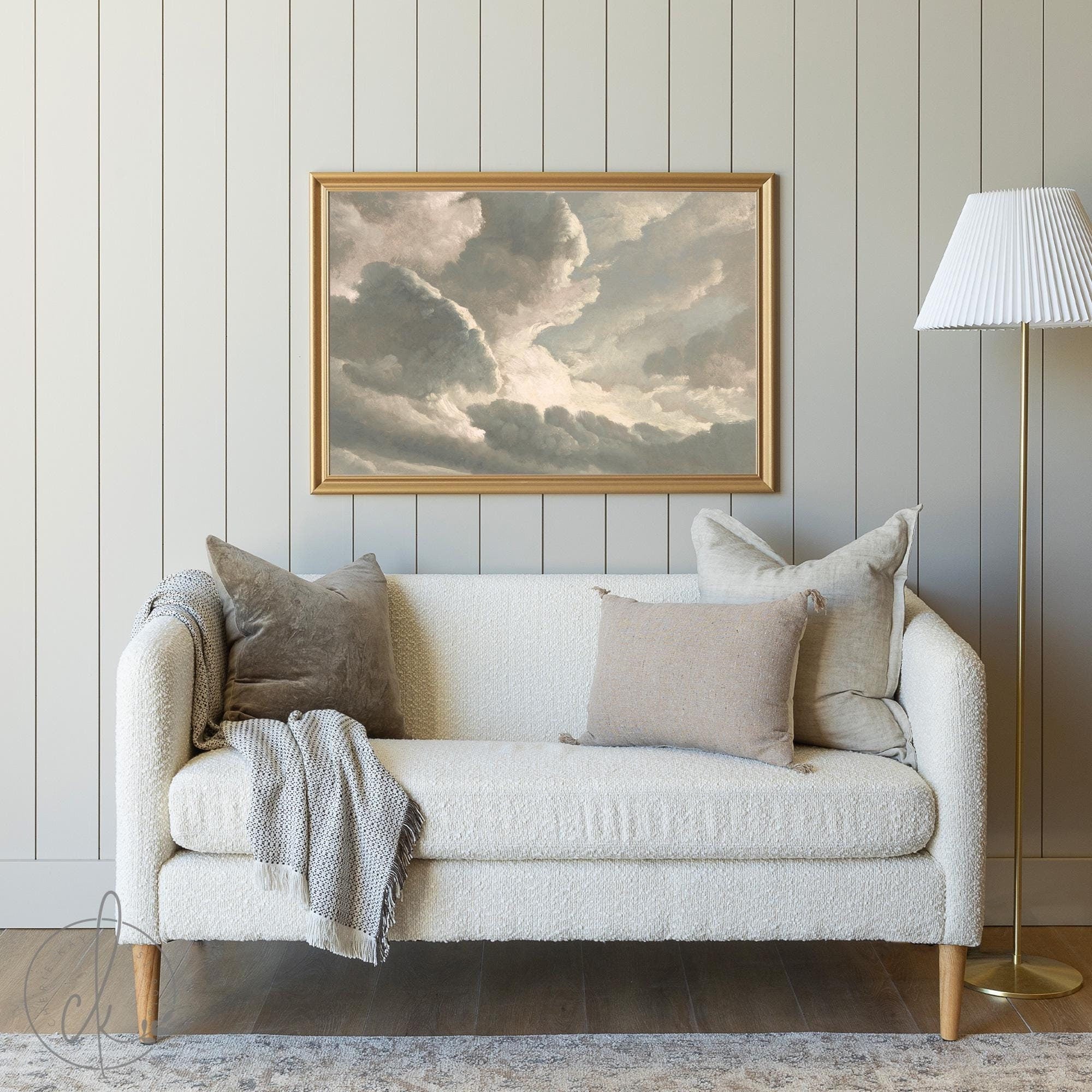 A cozy living room featuring a cream-colored sofa with neutral throw pillows and a blanket, set against a paneled wall. A framed painting of illuminated clouds hangs above the sofa, complemented by a gold floor lamp with a white shade on the side.