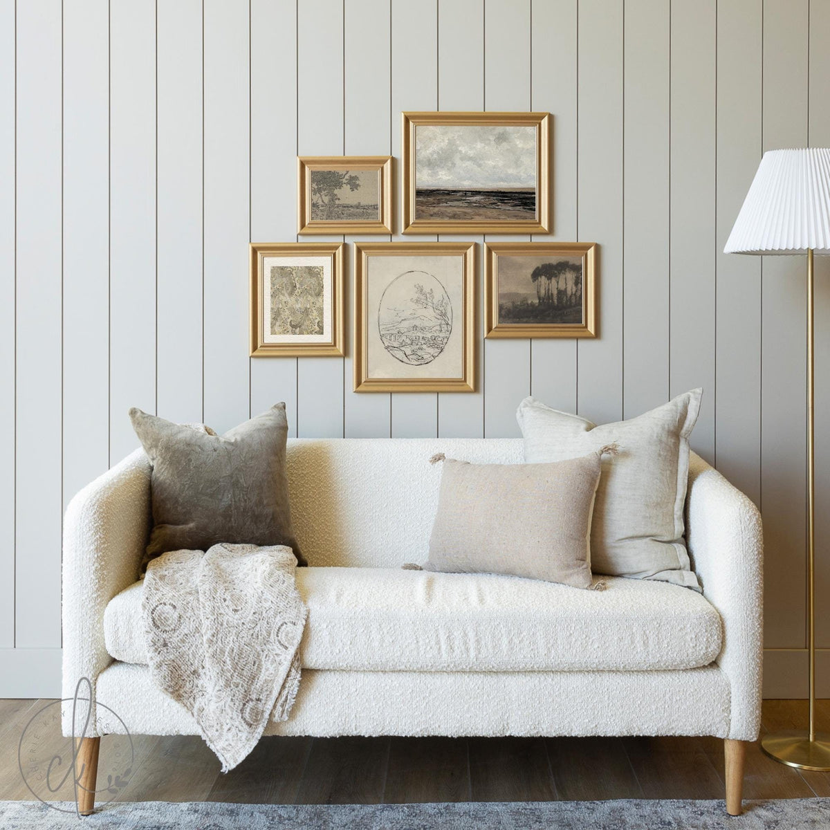 A cozy living room with a white sofa, neutral pillows, a draped blanket, and gold-framed art on a light gray paneled wall. A modern floor lamp stands to the right.